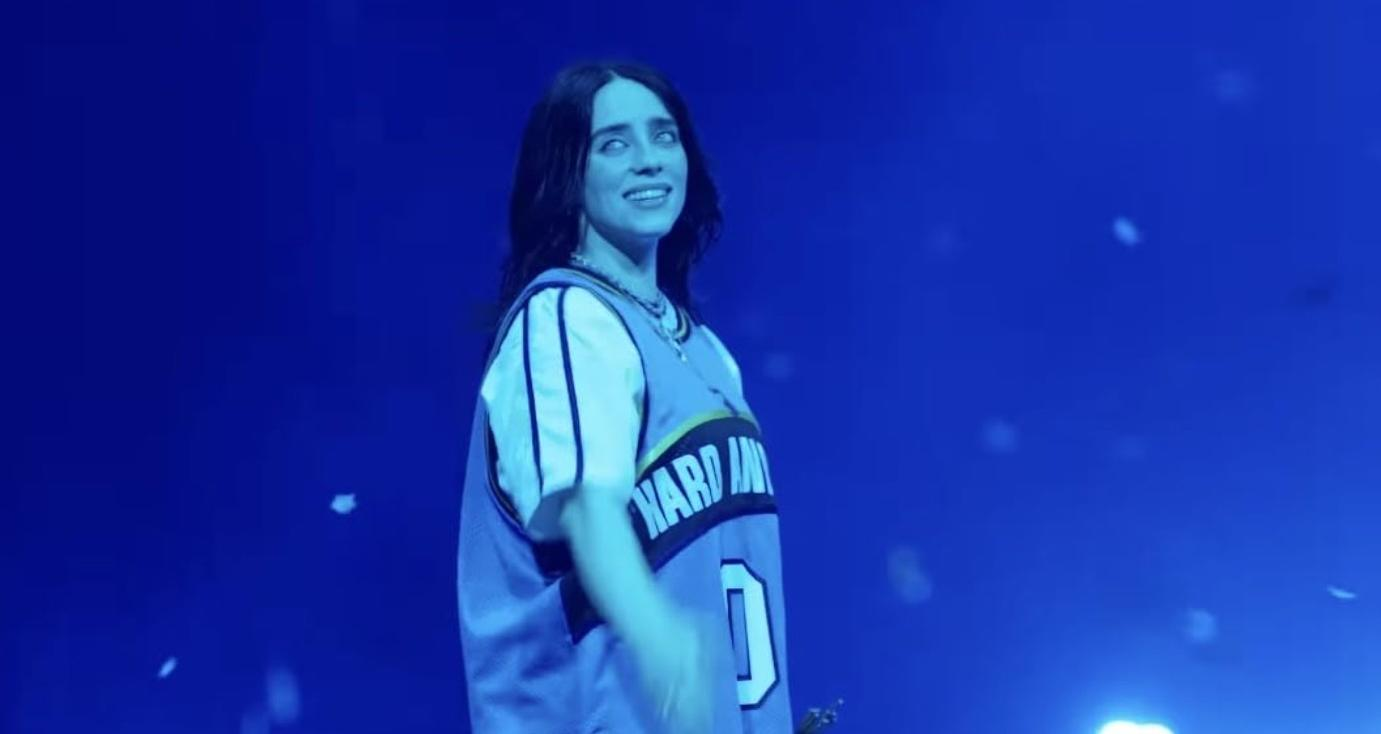 Eine Frau mit dunklen Haaren trägt ein hellblaues Trikot auf einer Bühne mit blauem Hintergrund.