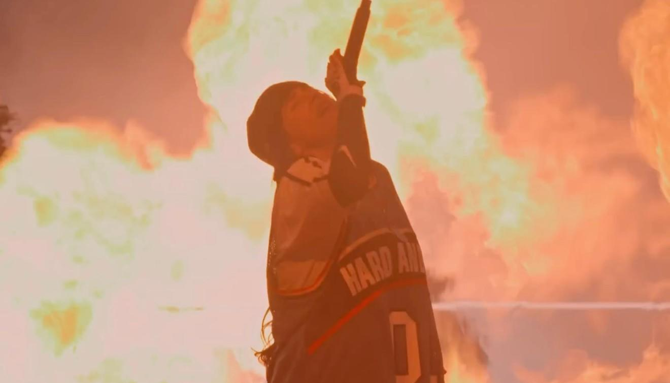 Eine Person mit einem Mikrofon vor orangefarbenen Flammen.