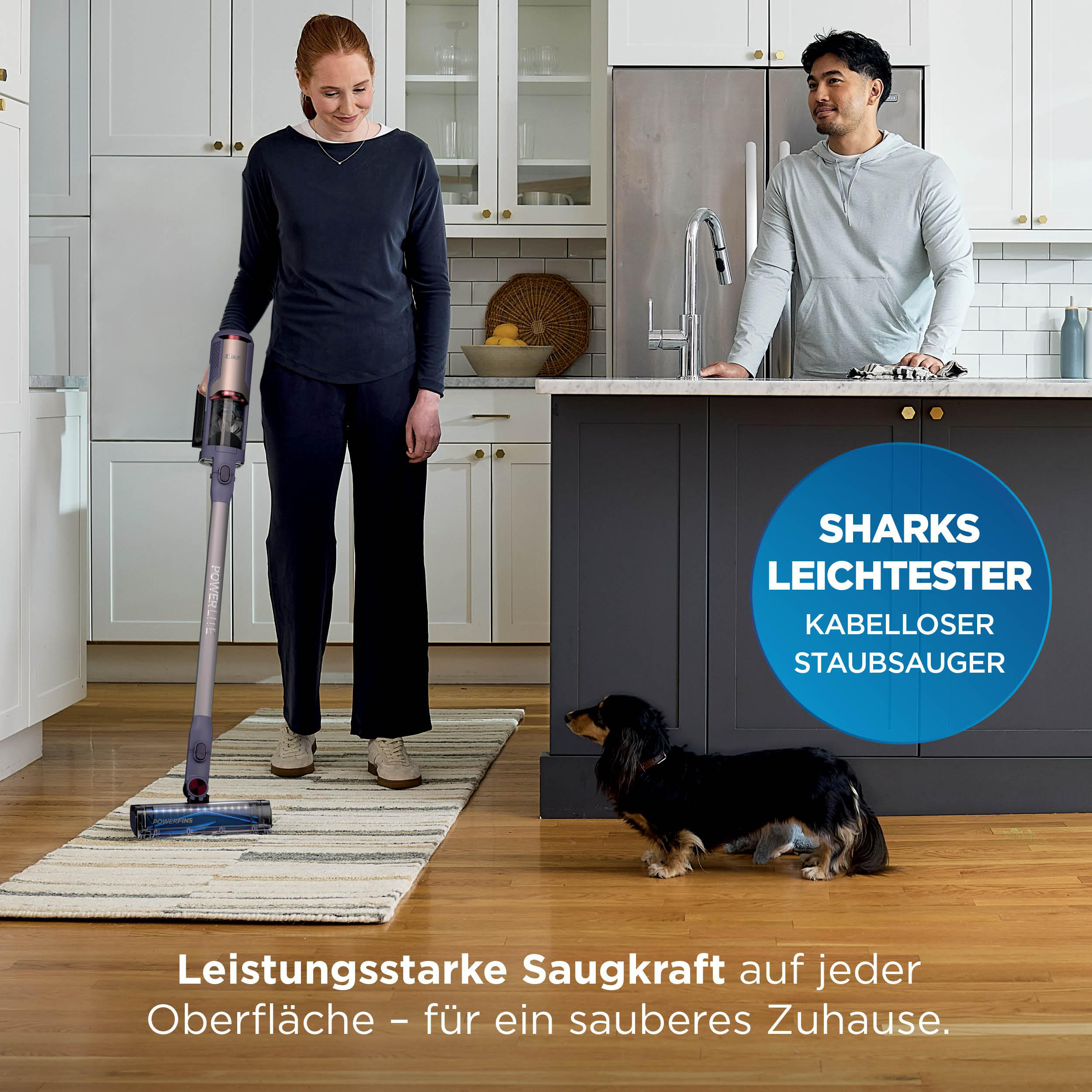 Frau saugt Küche mit Akkusauger, Mann steht daneben, Hund wartet.