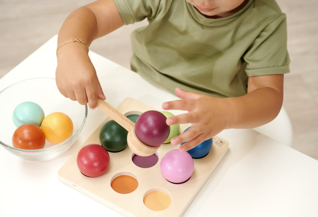 Ein Kind spielt mit bunten Holzklötzen auf einem Brett. Ein Löffel hält einen violetten Klotz.