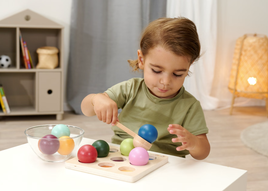 Ein Kleinkind mit einem Löffel spielt mit bunten Kugeln auf einem Holzbrett.