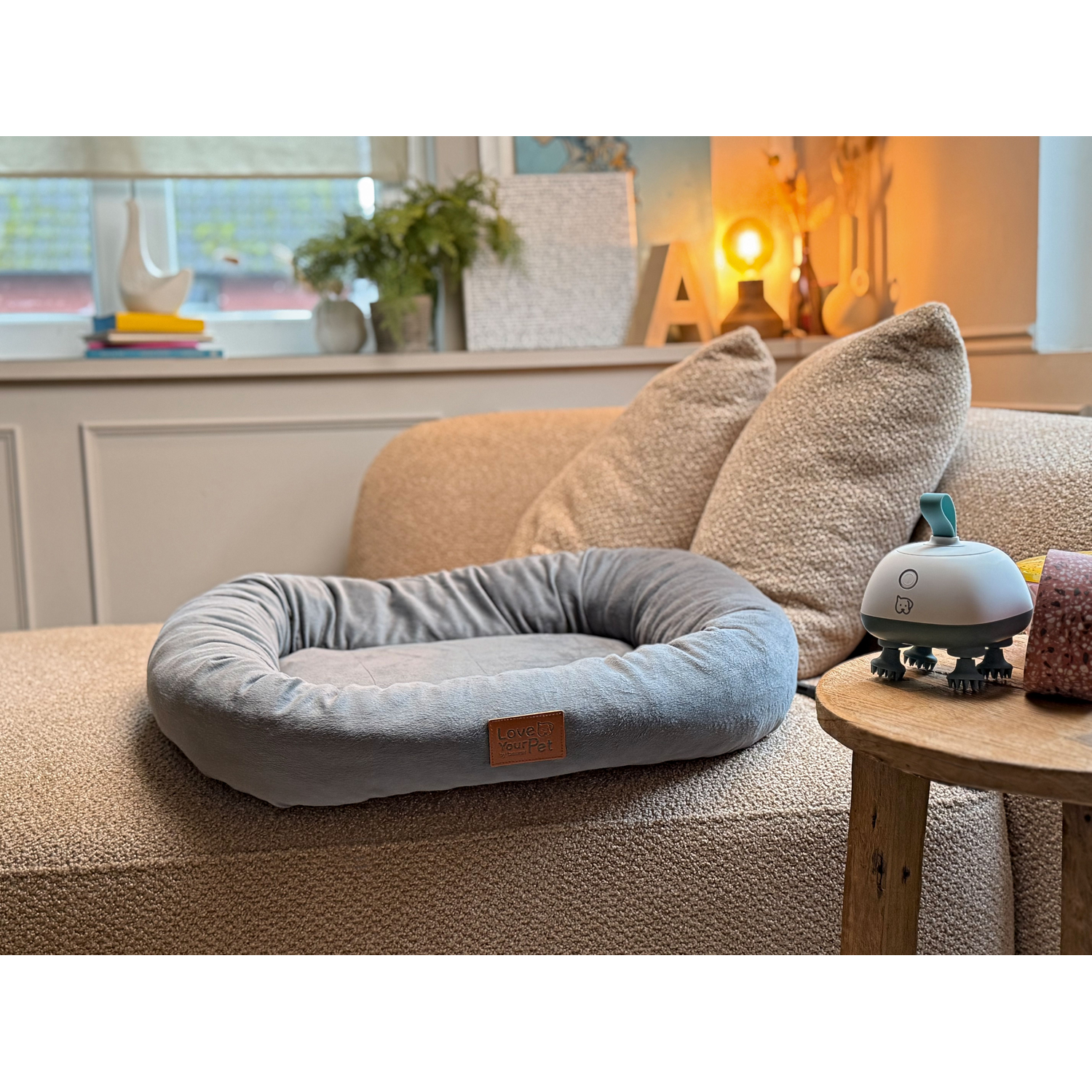 Een grijs pluche dierenbed staat op een stoffen bank naast een klein houten tafeltje met een dierenmassager.