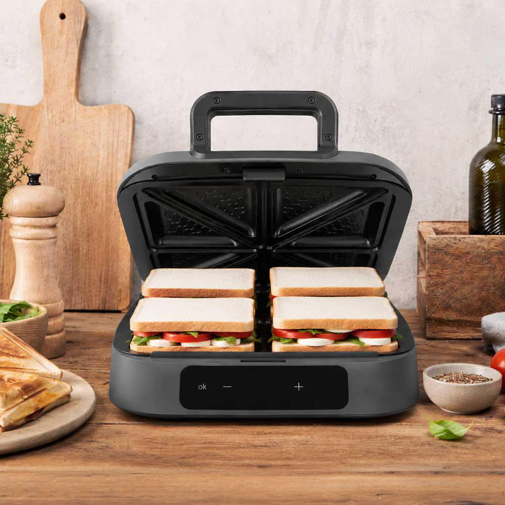 Ein offener Sandwichmaker mit vier Sandwiches, bereit zum Toasten, auf einem Holztisch.