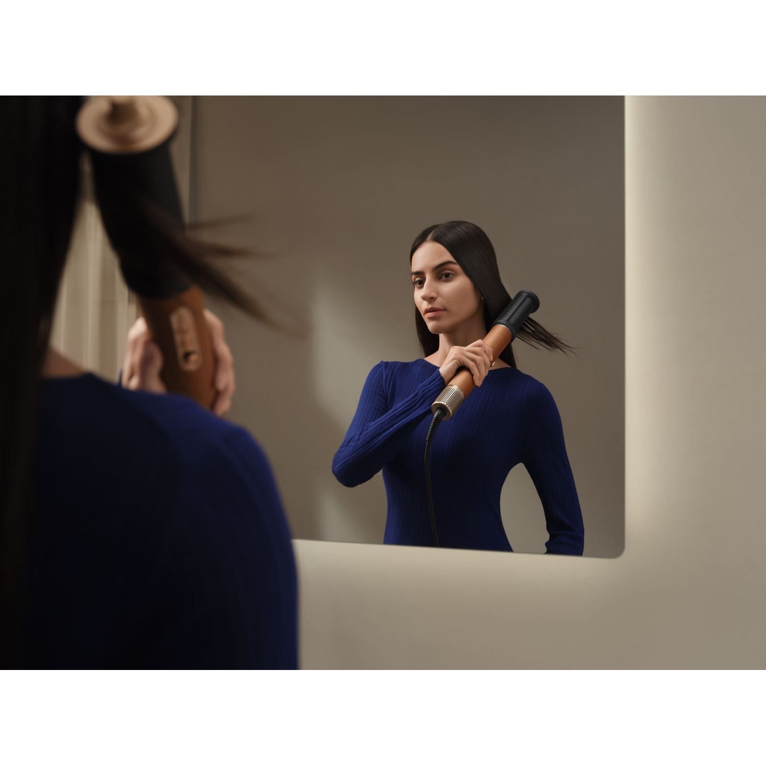A woman with long dark hair styles her hair using a hair dryer.