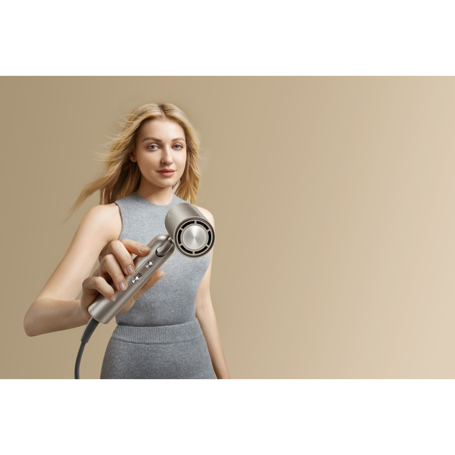 A blonde woman holds a silver hairdryer in front of a beige background.