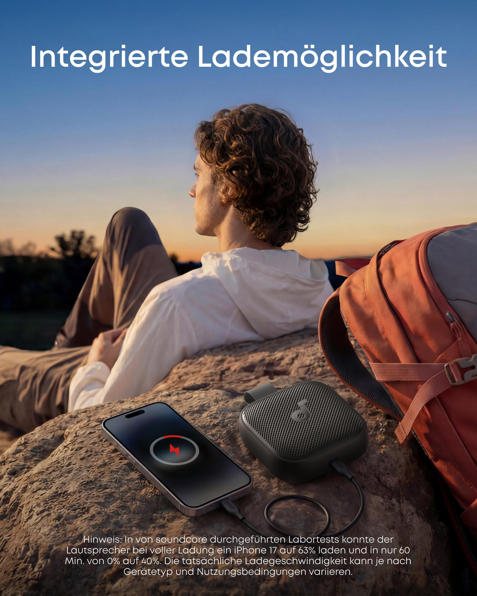 Mann im Freien lädt Handy mit Lautsprecher, Rucksack nebenan.