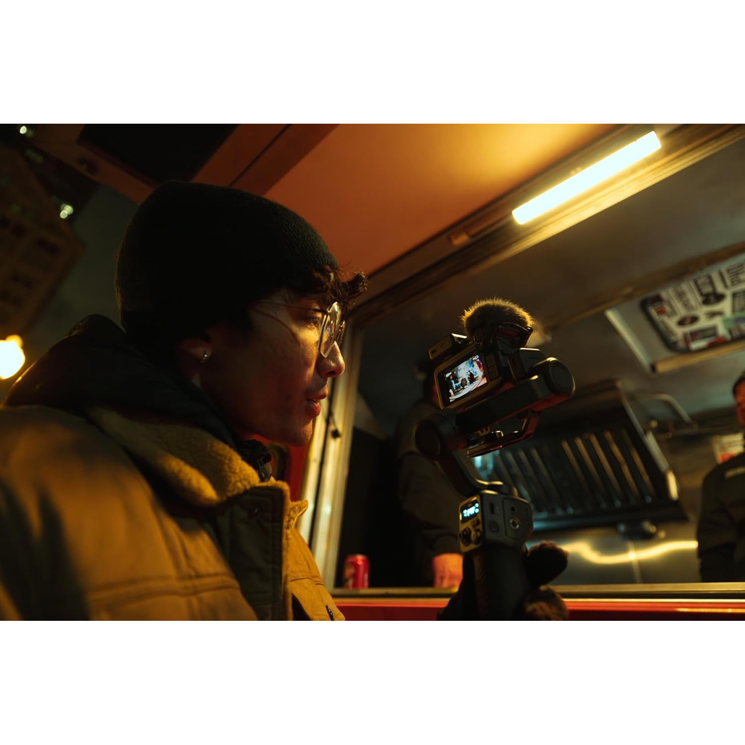 Person with glasses and beanie films food truck with gimbal camera at night.