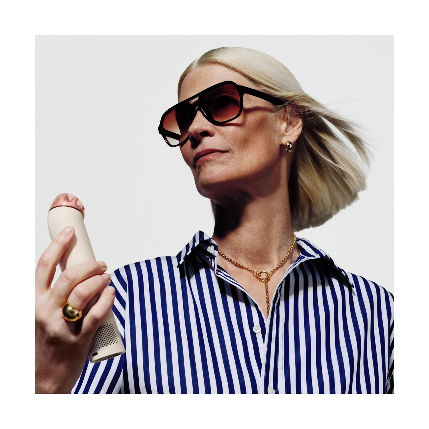 Donna con capelli biondi, occhiali da sole e camicia a righe blu tiene un dispositivo.