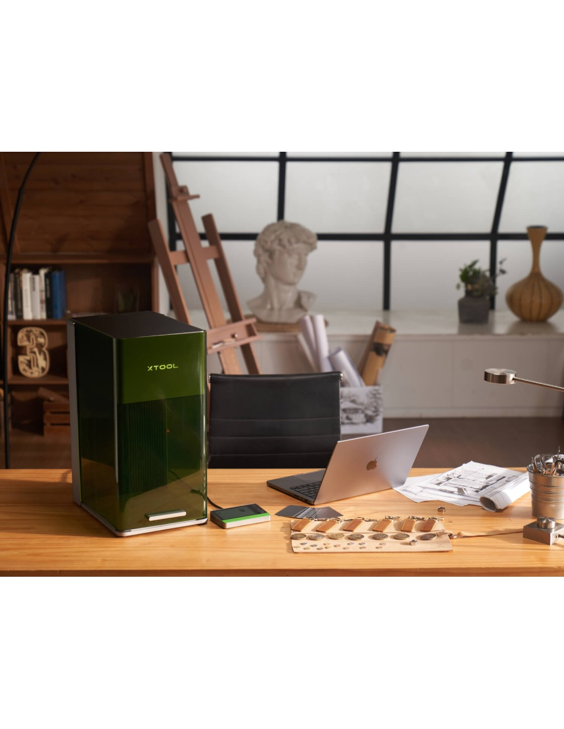 Un dispositivo verde está sobre un escritorio de madera junto a un portátil, planos y un busto.