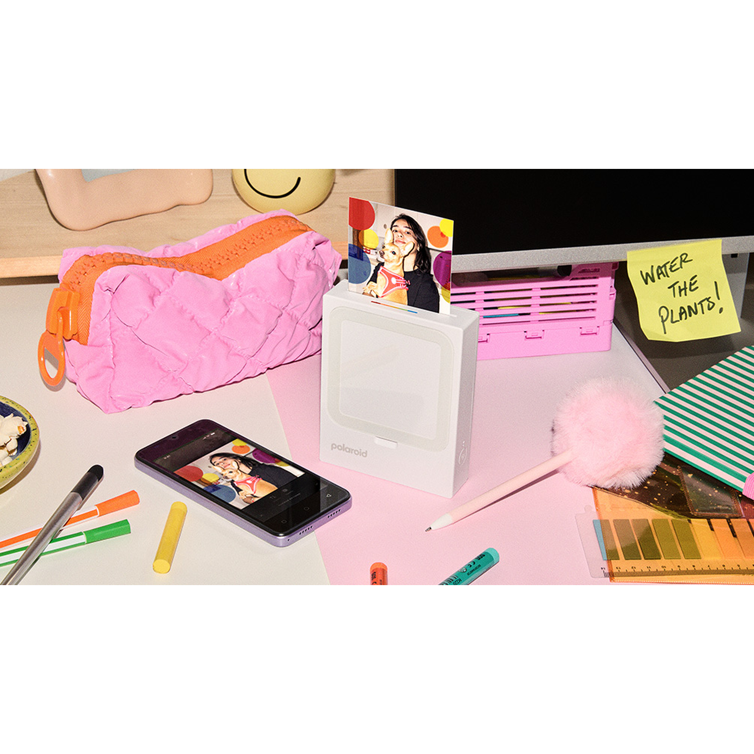 A desk with a Polaroid printer, phone, pens, and a pink bag.