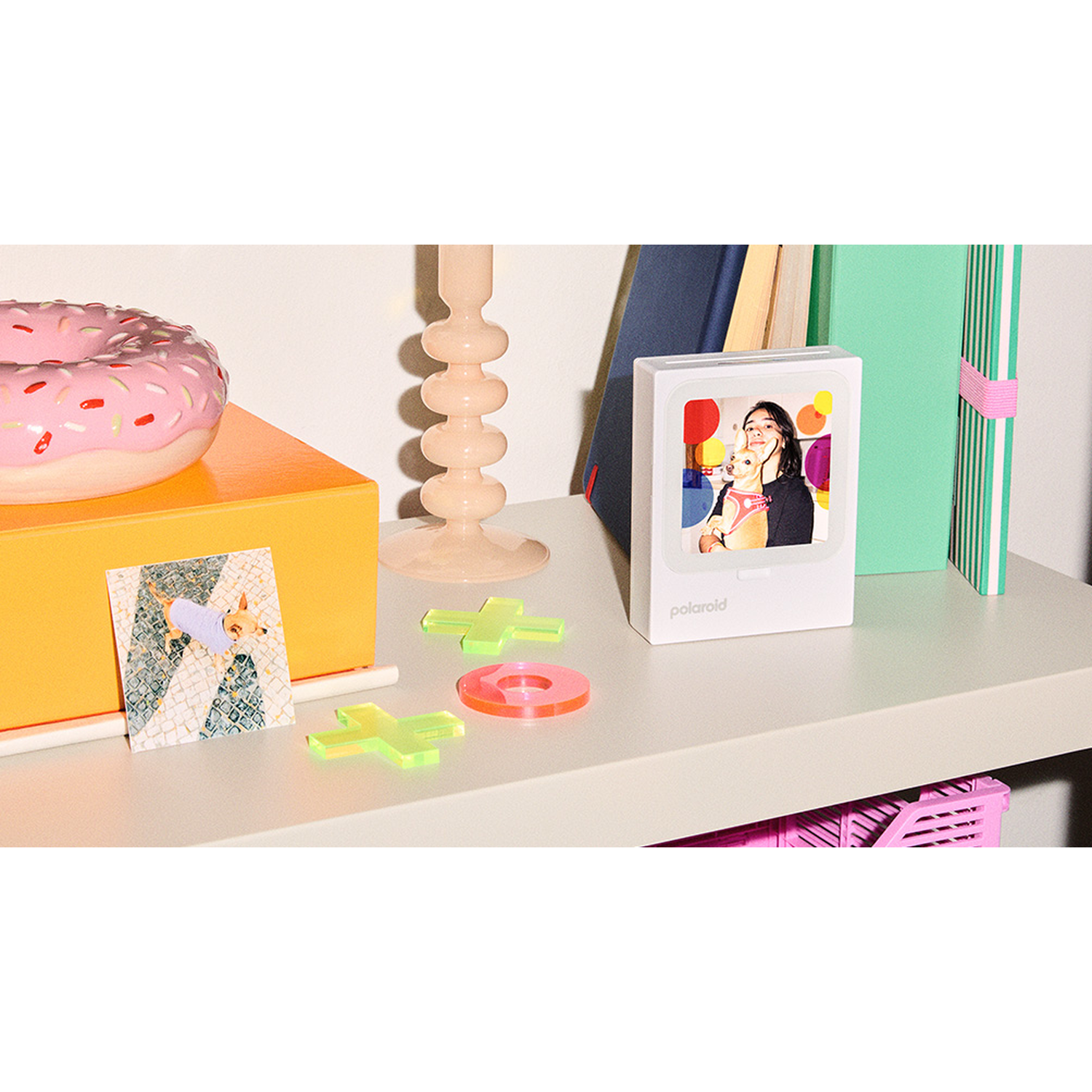 A Polaroid printer with a photo, donut, candle, books, and decorations on a shelf.