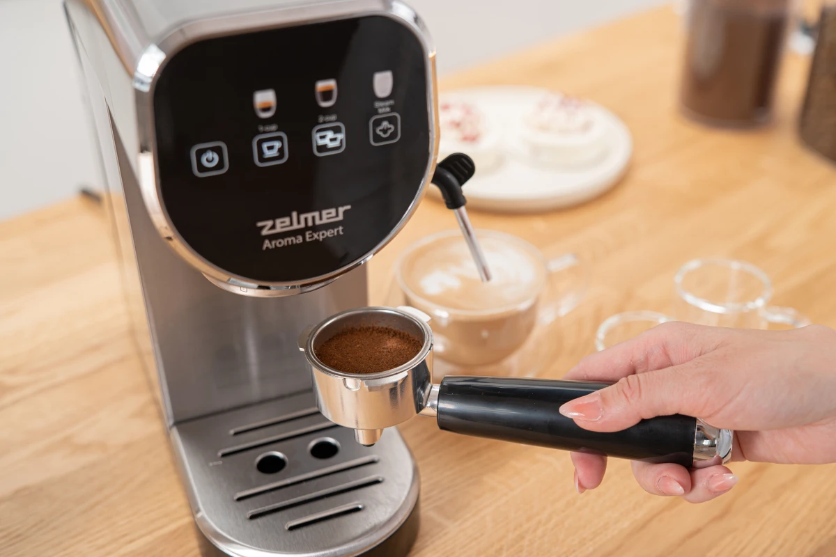Dłoń trzyma uchwyt filtra z kawą mieloną obok ekspresu Zelmer Aroma Expert.