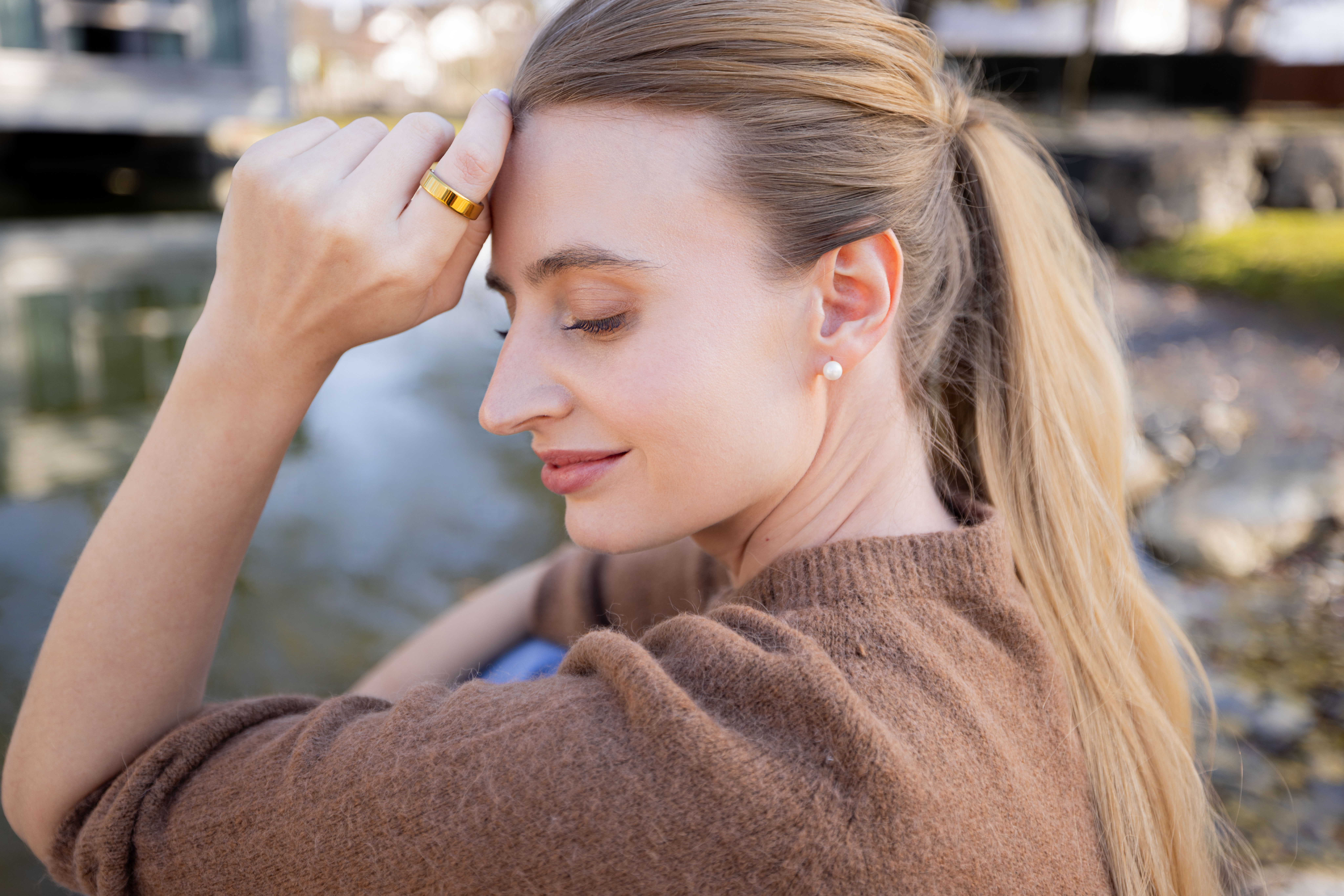 Frau mit blondem Haar, die einen braunen Pullover trägt, sitzt am Wasser.