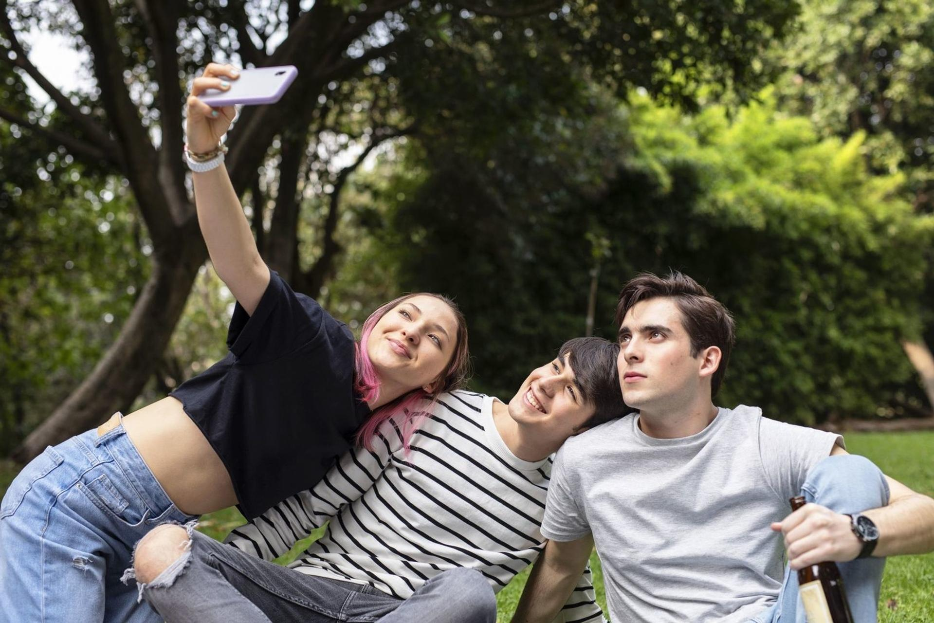 Drei junge Leute machen ein Selfie im Park. Eine Frau mit pinken Haaren hält das Handy.