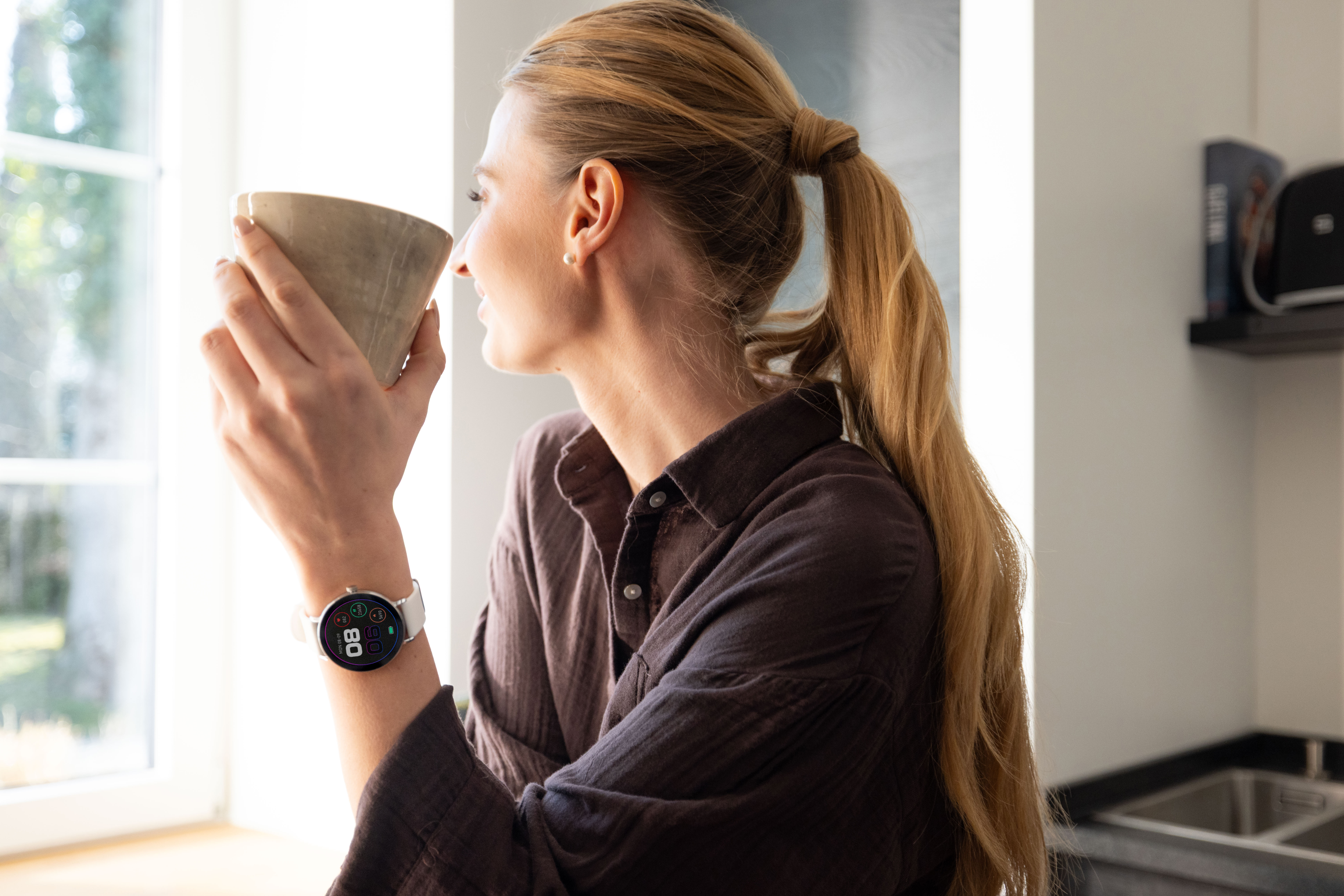 Frau mit Pferdeschwanz trinkt aus einer Tasse, trägt eine Smartwatch.