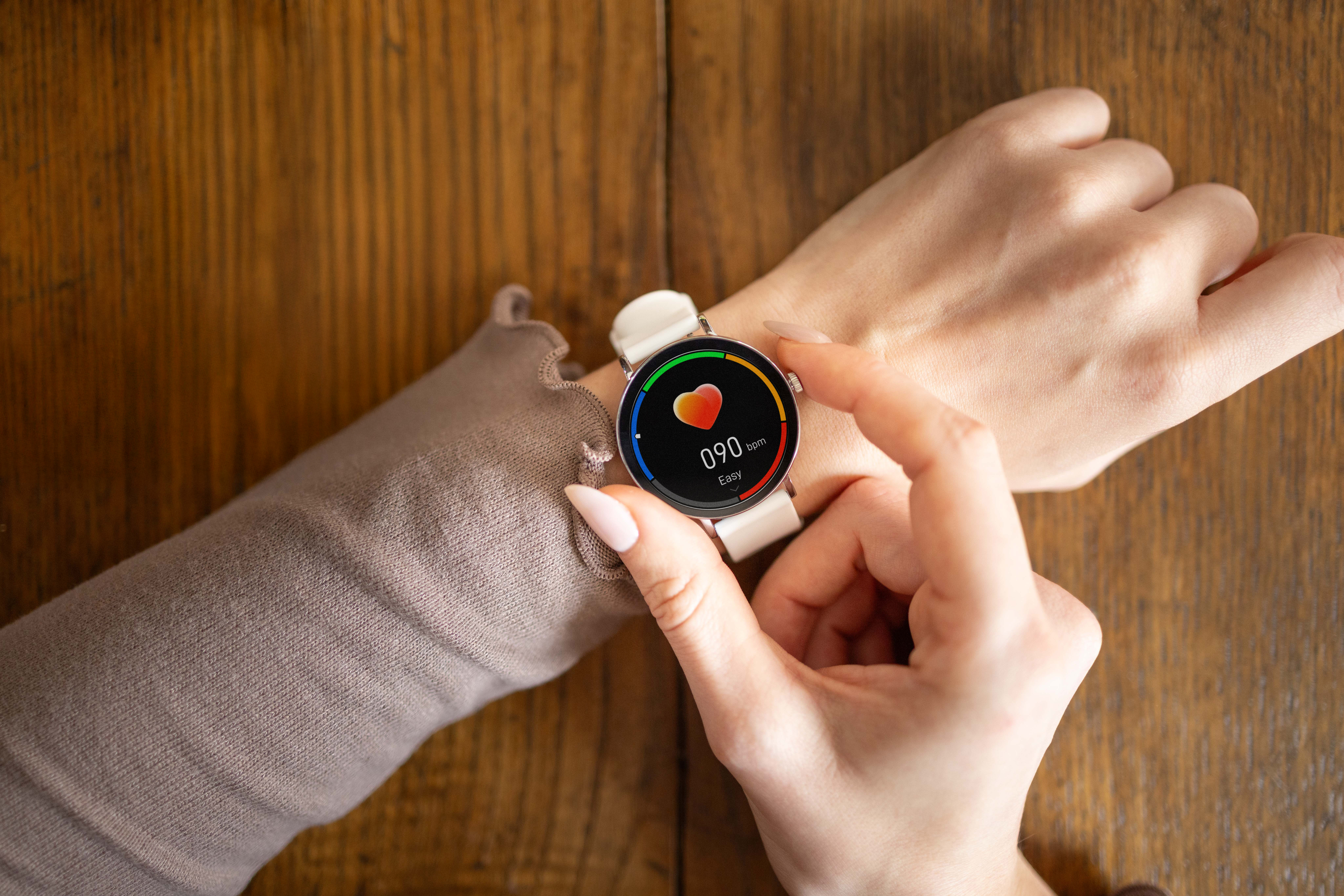 Eine Hand mit einem weißen Smartwatch-Armband, die einen Bildschirm mit Herzfrequenz anzeigt.