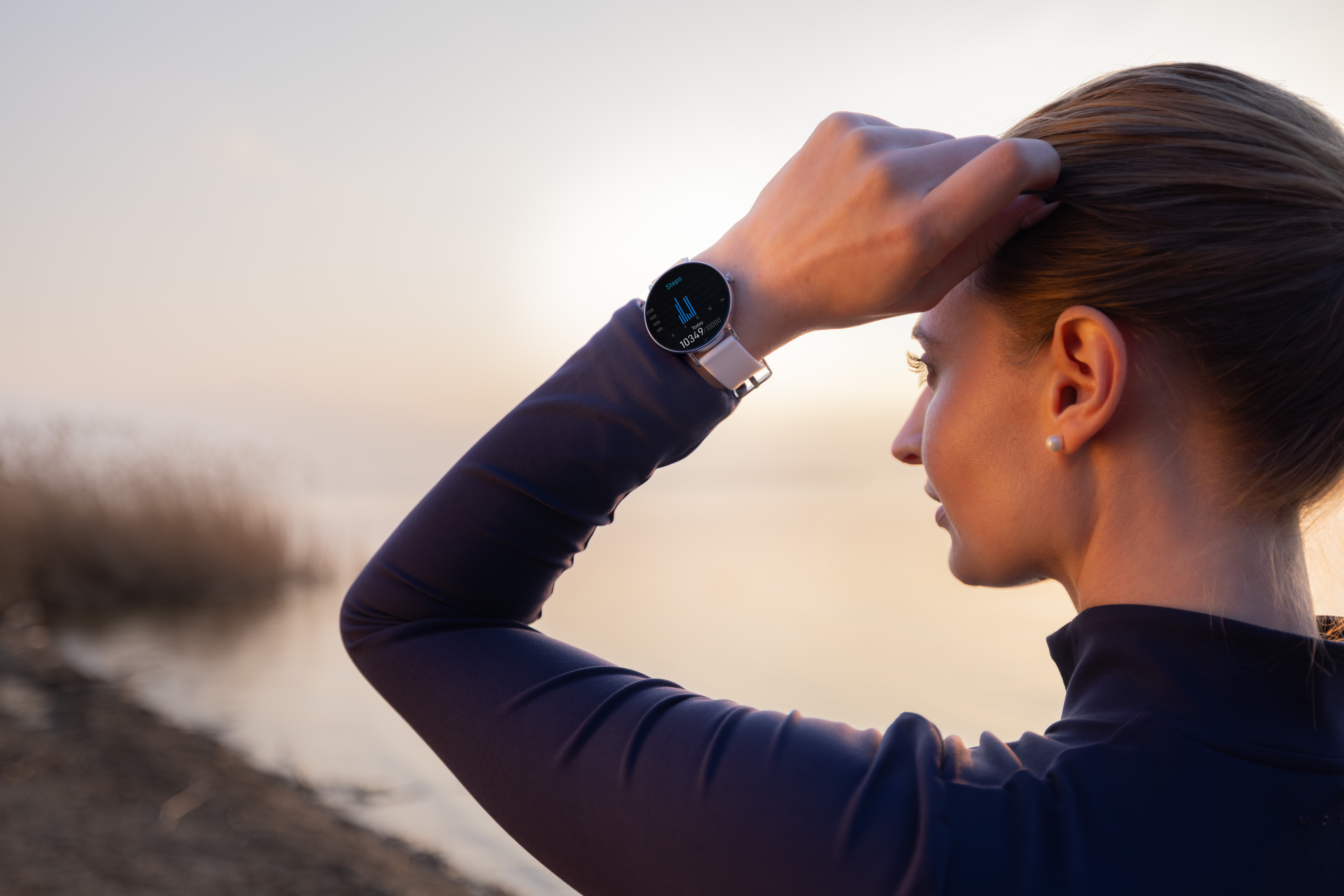 Frau mit Uhr am Handgelenk vor einem nebligen See bei Sonnenaufgang.