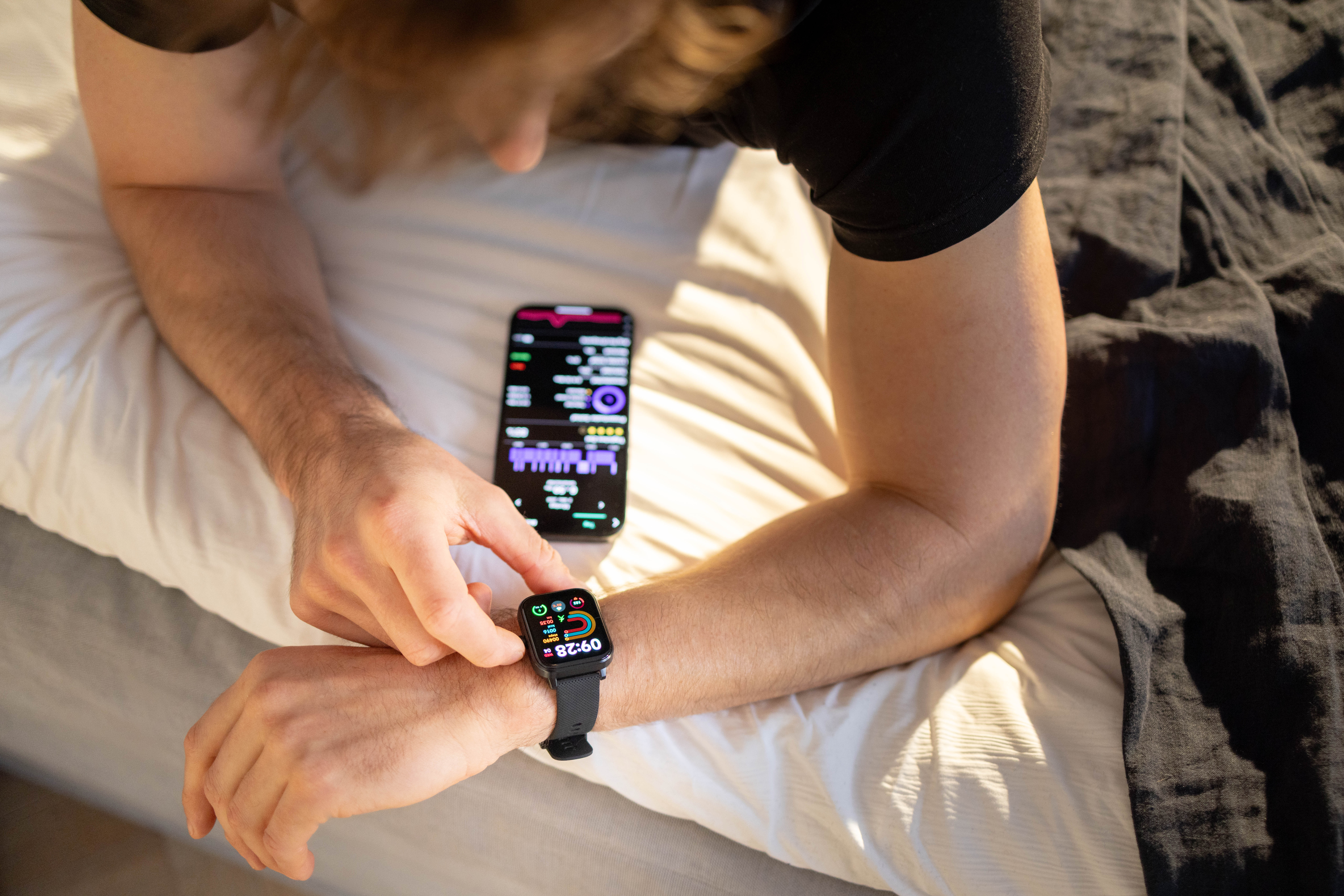 Eine Person prüft eine Smartwatch und ein Handy auf einem Bett mit weißer und grauer Bettwäsche.