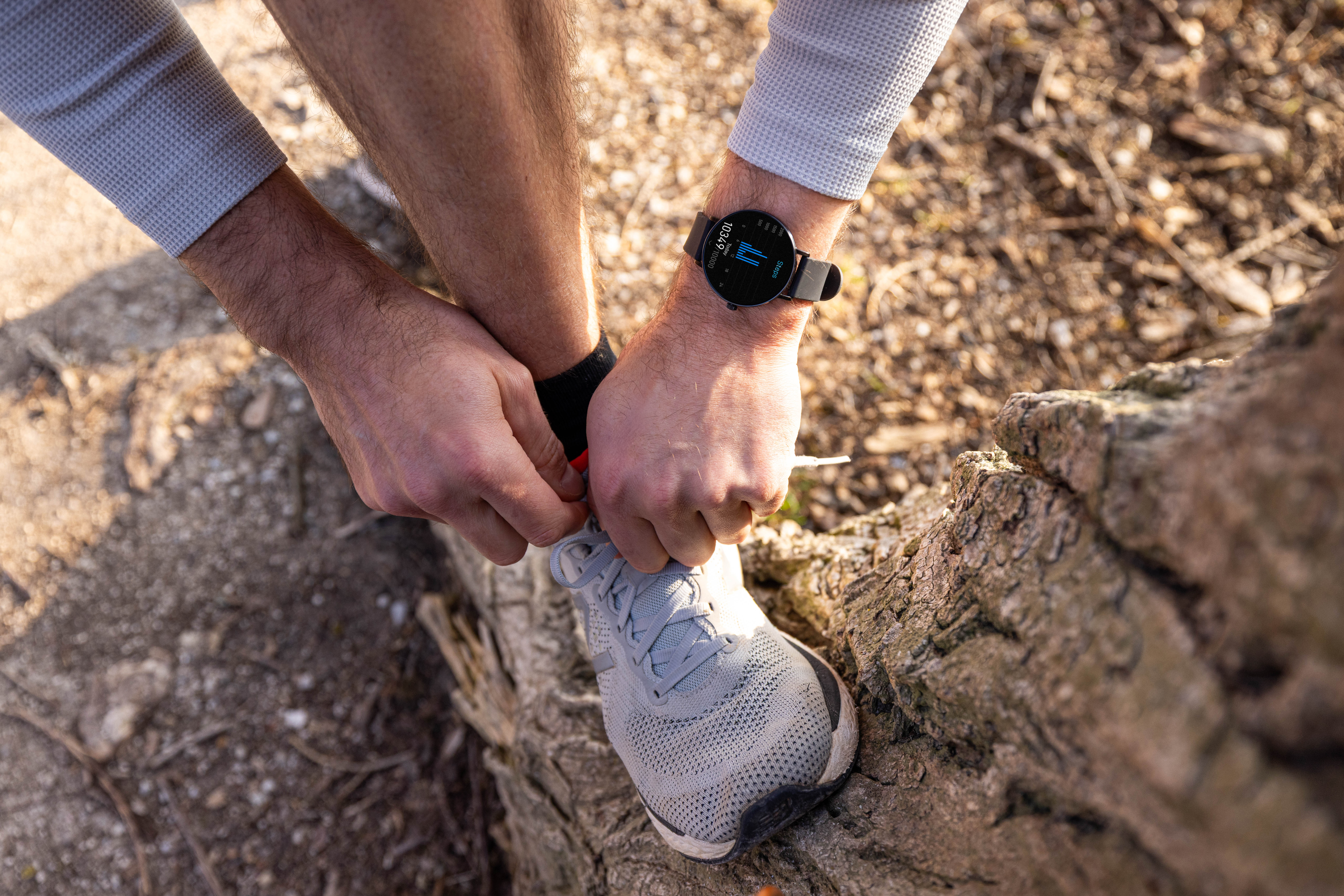 Mann bindet graue Laufschuhe auf steinigem Untergrund, trägt Smartwatch.