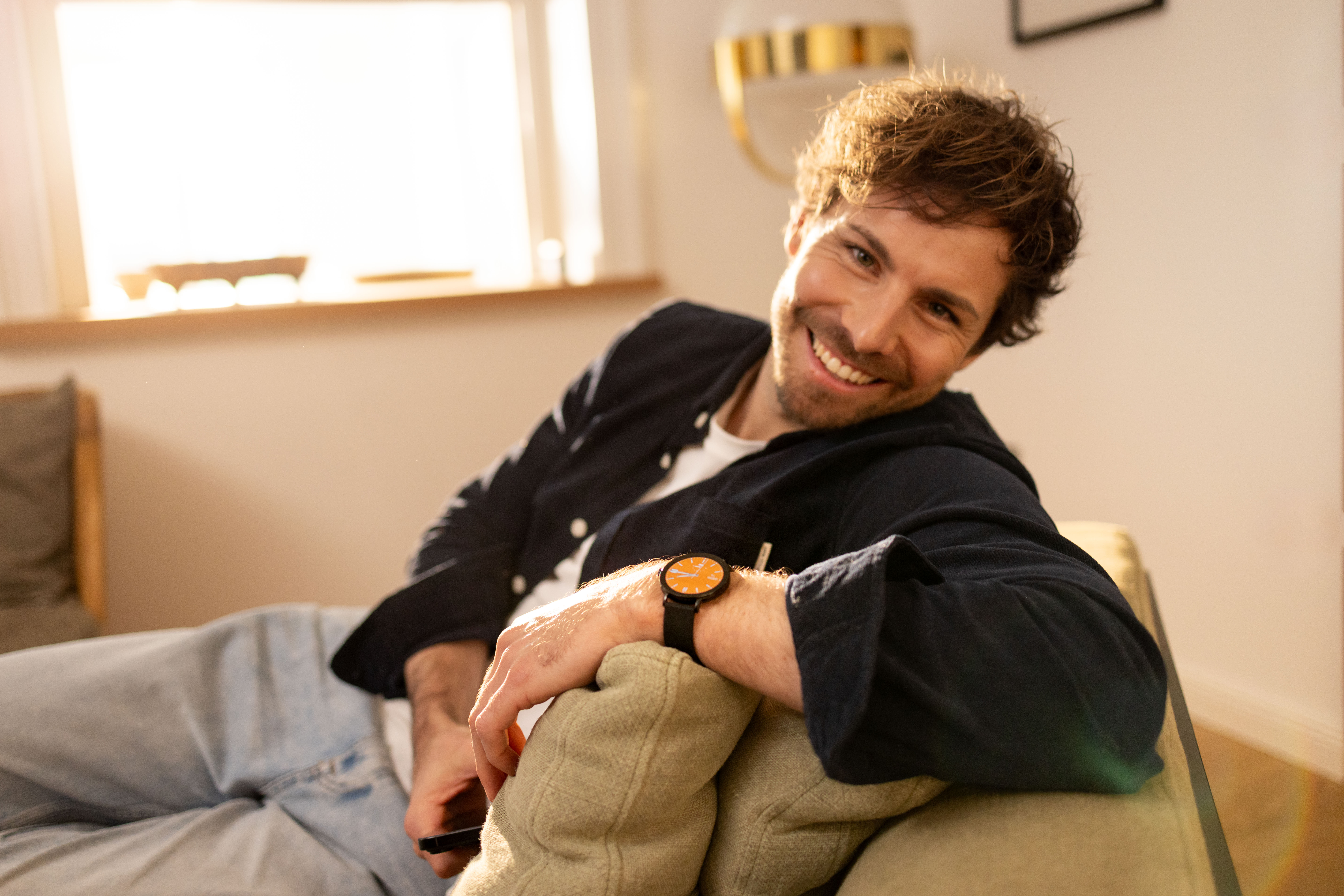 Ein Mann mit braunen Haaren sitzt auf einer Couch und lächelt. Er trägt ein dunkles Hemd.