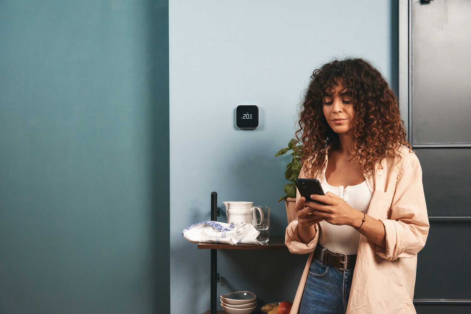 Mujer con pelo rizado usa smartphone frente a termostato de pared azul.