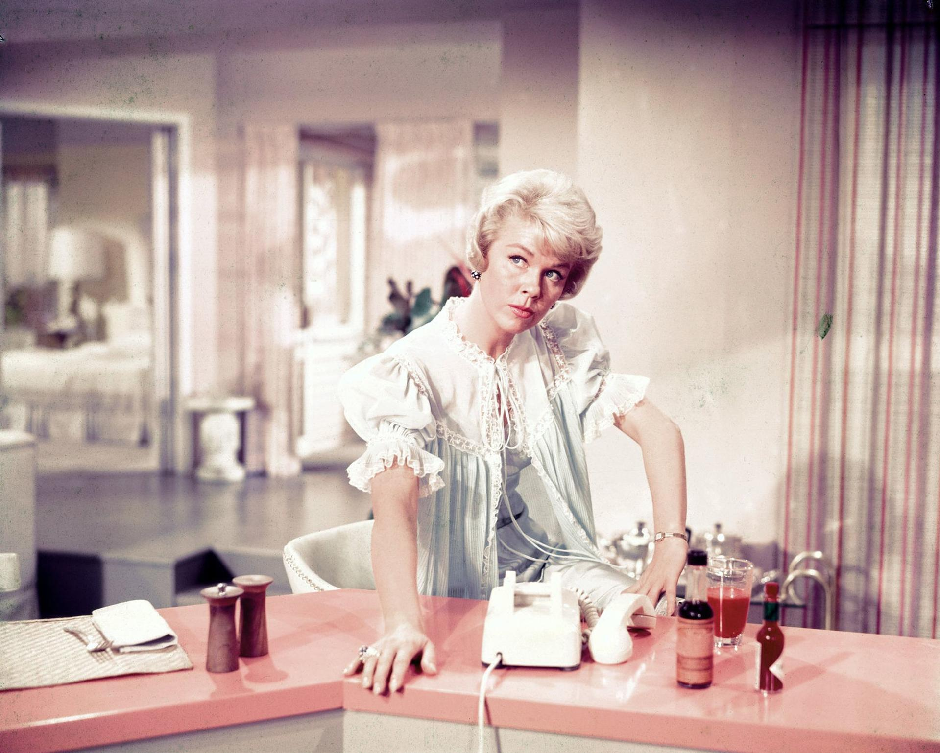 Eine blonde Frau in einem hellblauen Morgenmantel sitzt an einer rosa Theke mit einem weißen Wähltelefon, Salz- und Pfefferstreuern und Getränken.