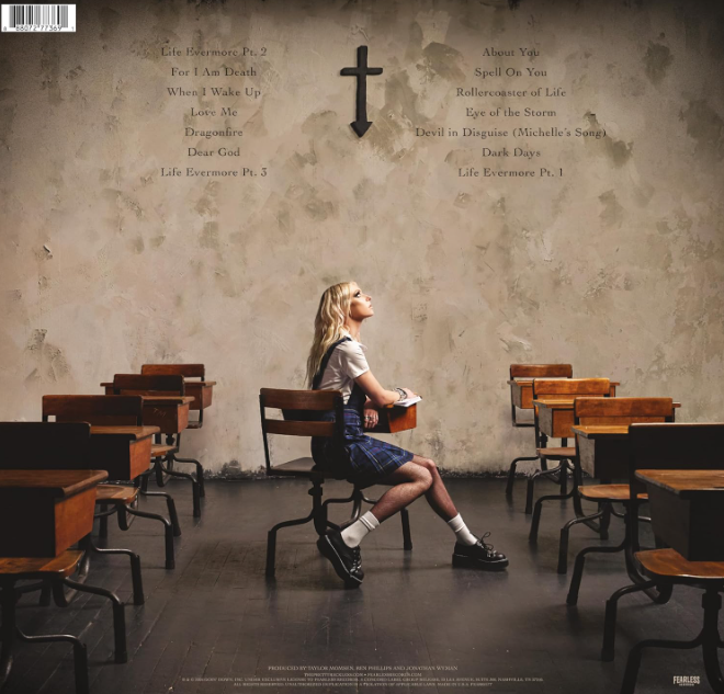 Eine blonde Frau in einem karierten Rock sitzt in einem Klassenzimmer und blickt zu einem Kreuz an einer strukturierten Wand mit aufgelisteten Songtiteln auf.