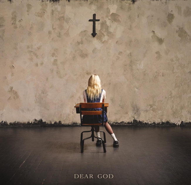 Eine blonde Person in Schuluniform sitzt an einem Schreibtisch und blickt auf eine strukturierte Wand mit einem schwarzen Kreuz darüber, darunter der Text „DEAR GOD“.