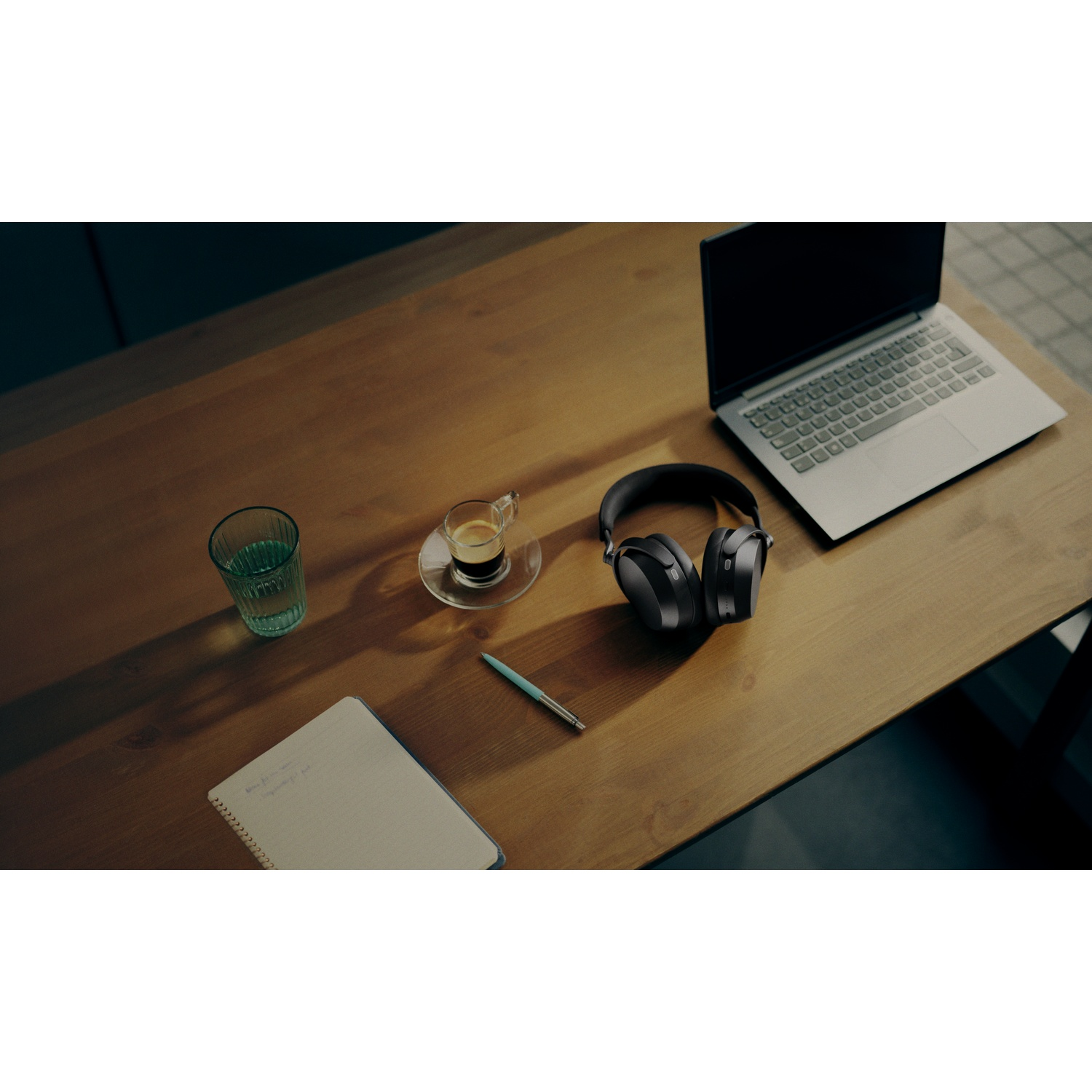 A desk with headphones, laptop, notebook, pen, and drinks.