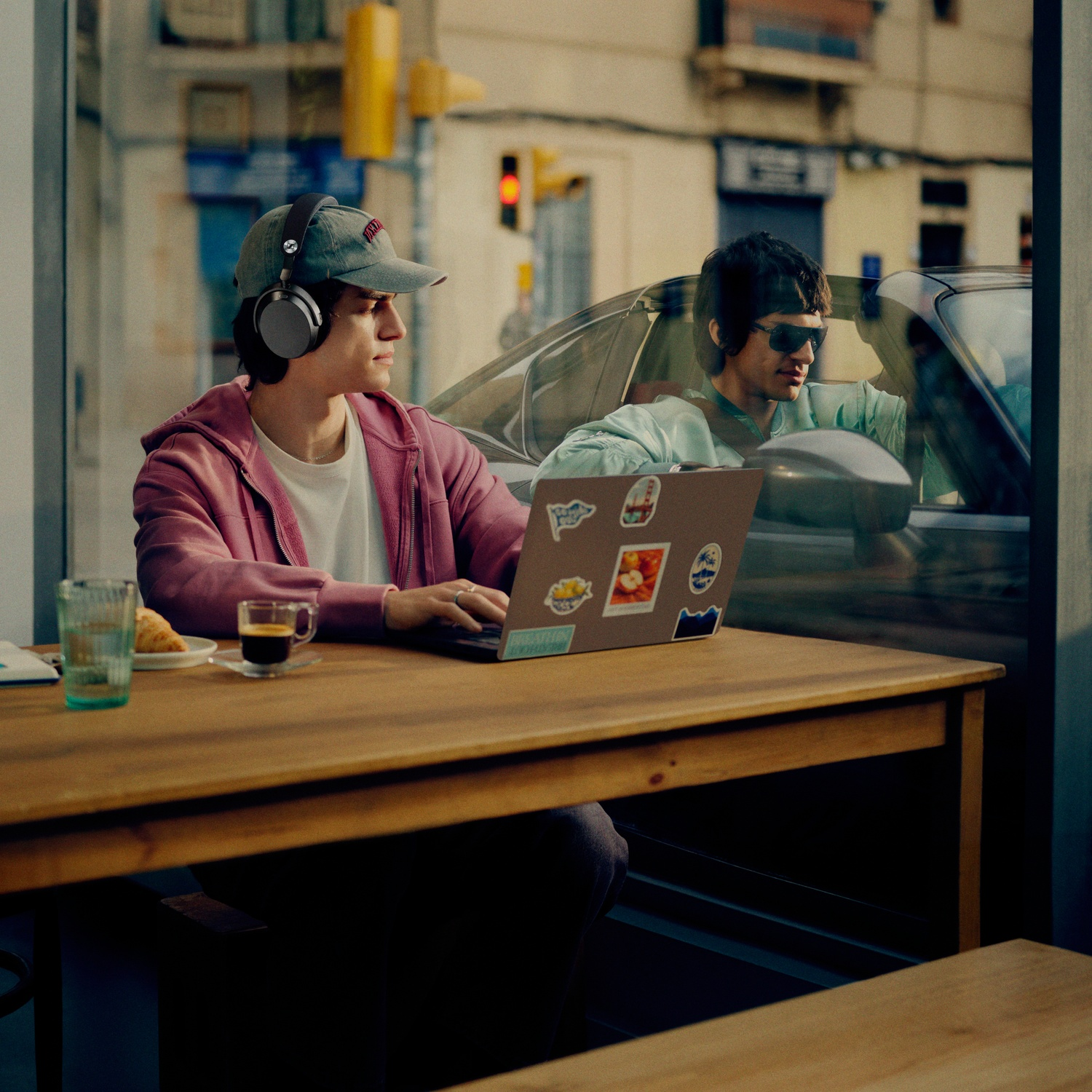 A man wearing headphones types on a laptop, while another man sits in a car.