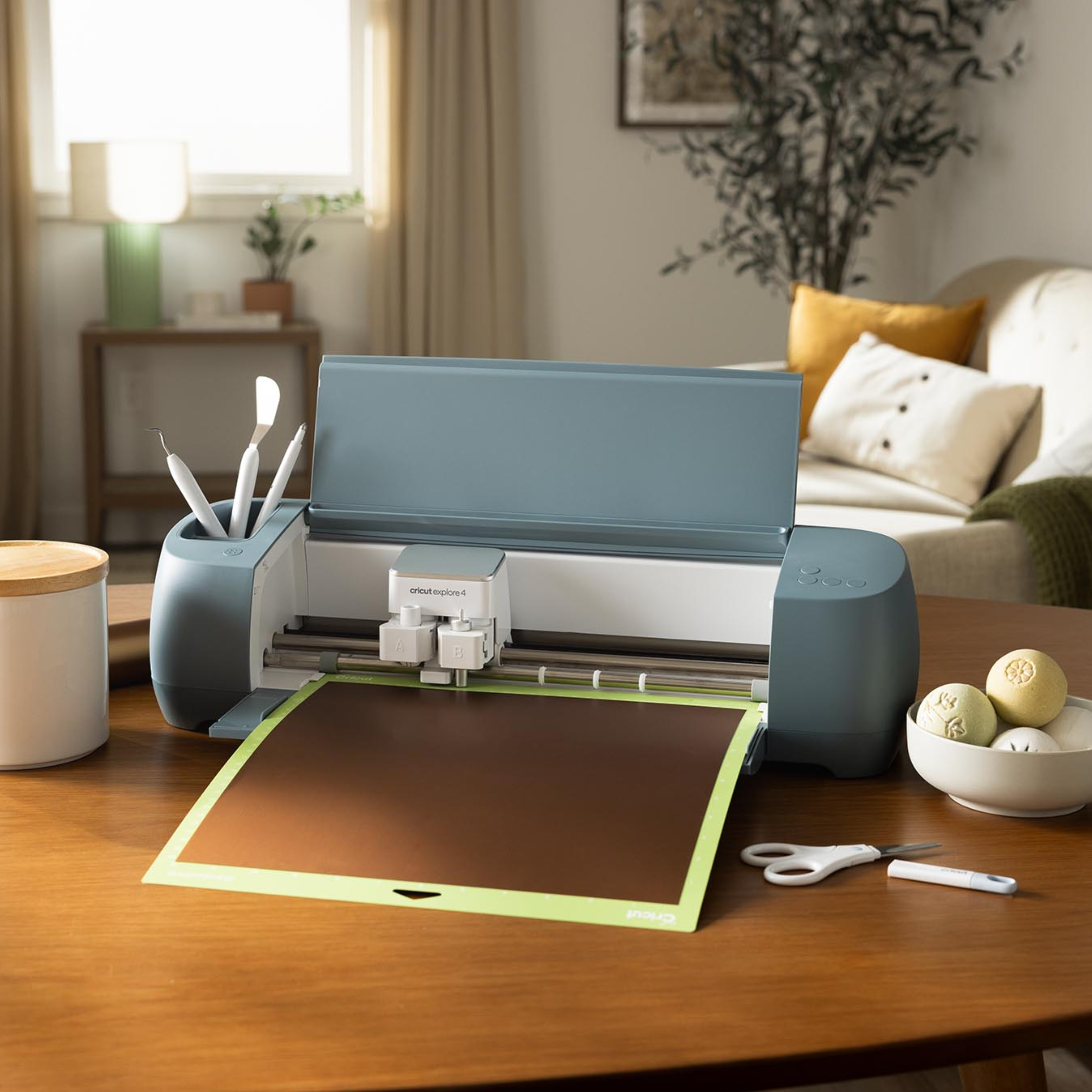 Mavi bir Cricut Explore Air 2 makinesi, aletler ve süslü topların olduğu bir kase ile ahşap bir masada duruyor.