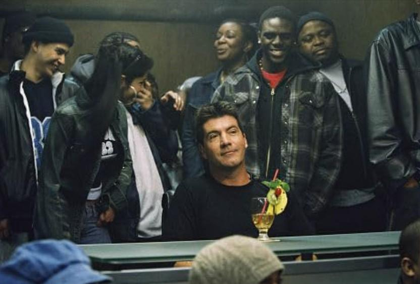 Simon Cowell sitzt mit einem Getränk, umgeben von Menschen. Hohe Dichte.