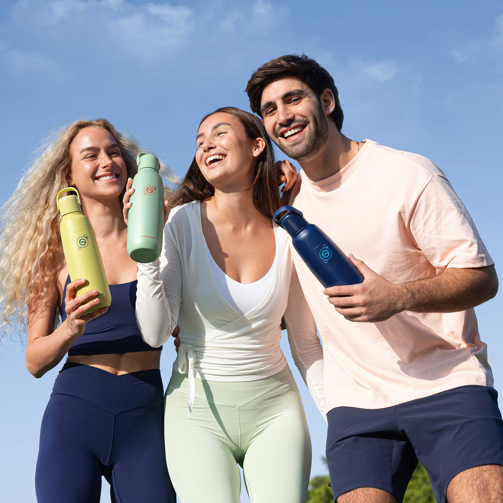 Három mosolygó ember sportruházatban színes, újrafelhasználható kulacsokat tart a szabadban.