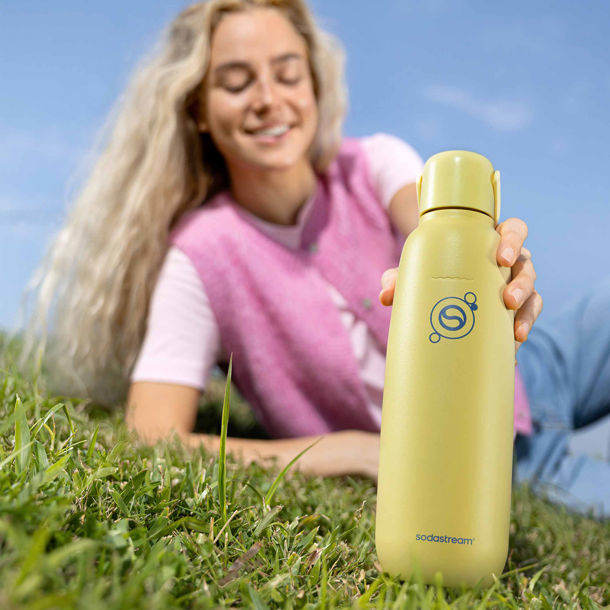 Egy nő a fűben fekszik, egy sárga Sodastream palackot tartva.