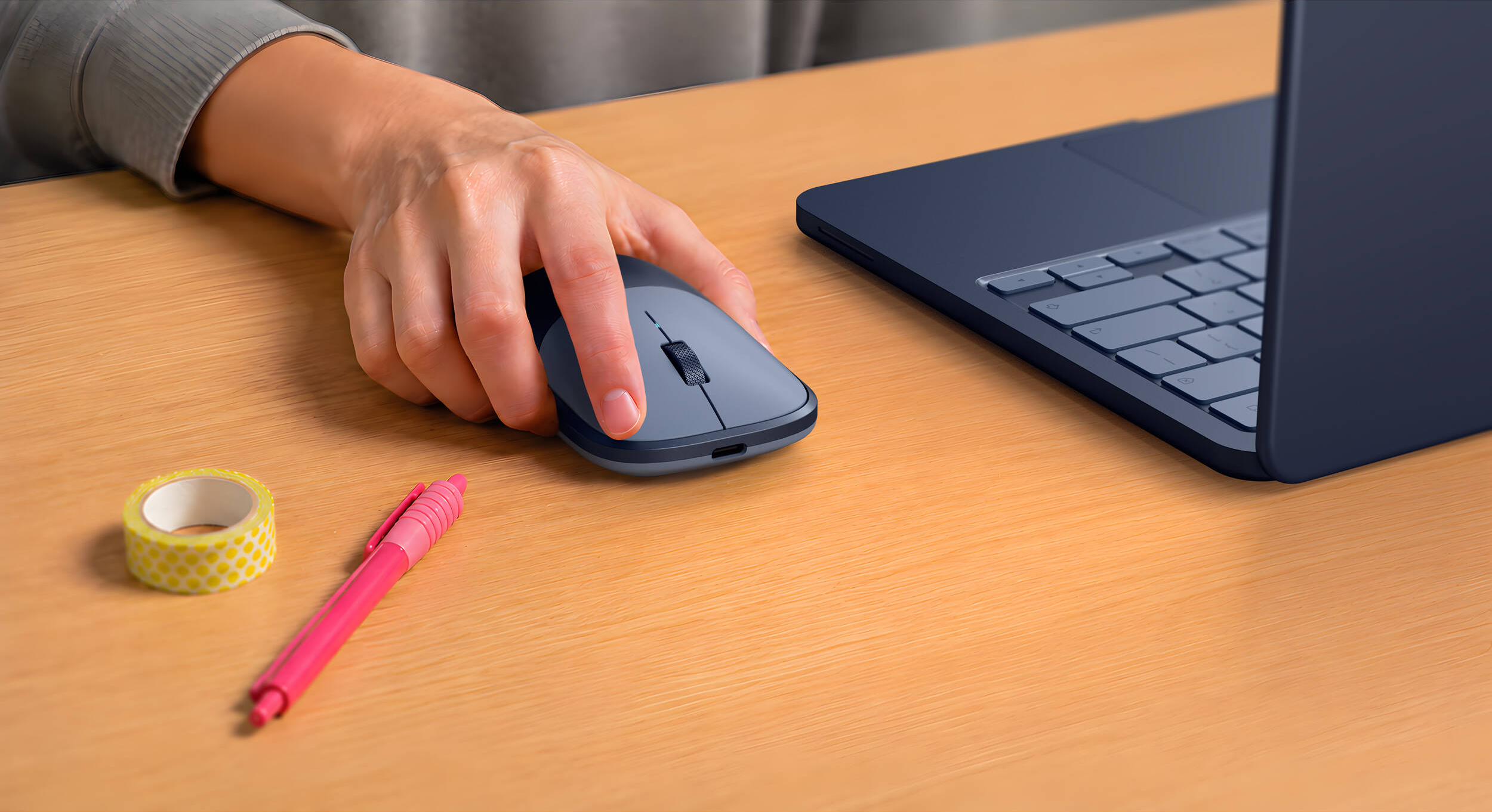 Une main utilise une souris grise à côté d'un ordinateur portable bleu foncé sur un bureau en bois.