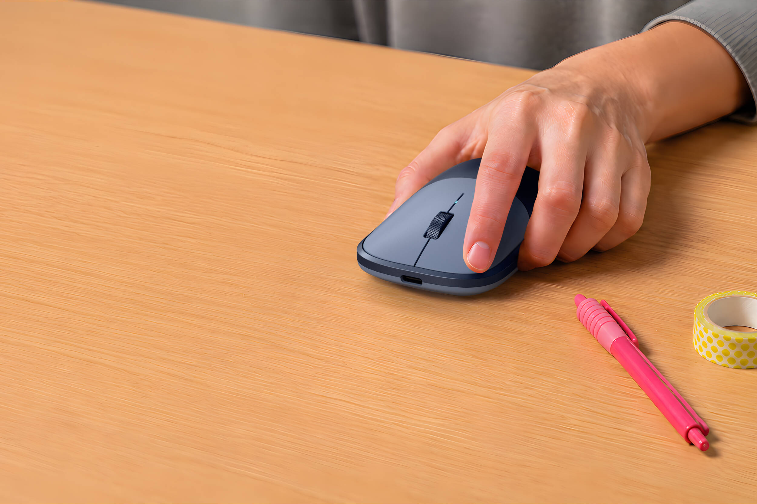Une main utilise une souris grise sur un bureau en bois. Un stylo rose et du ruban adhésif sont à côté.