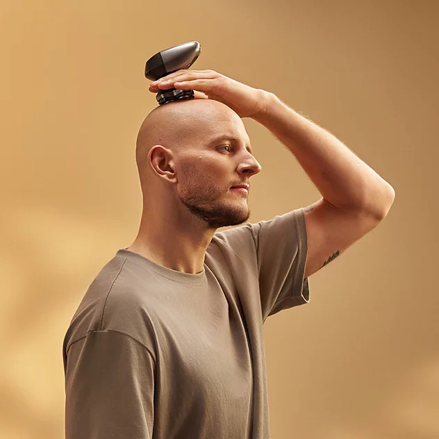 Łysy mężczyzna w szarym t-shircie trzyma czarną golarkę elektryczną na głowie.