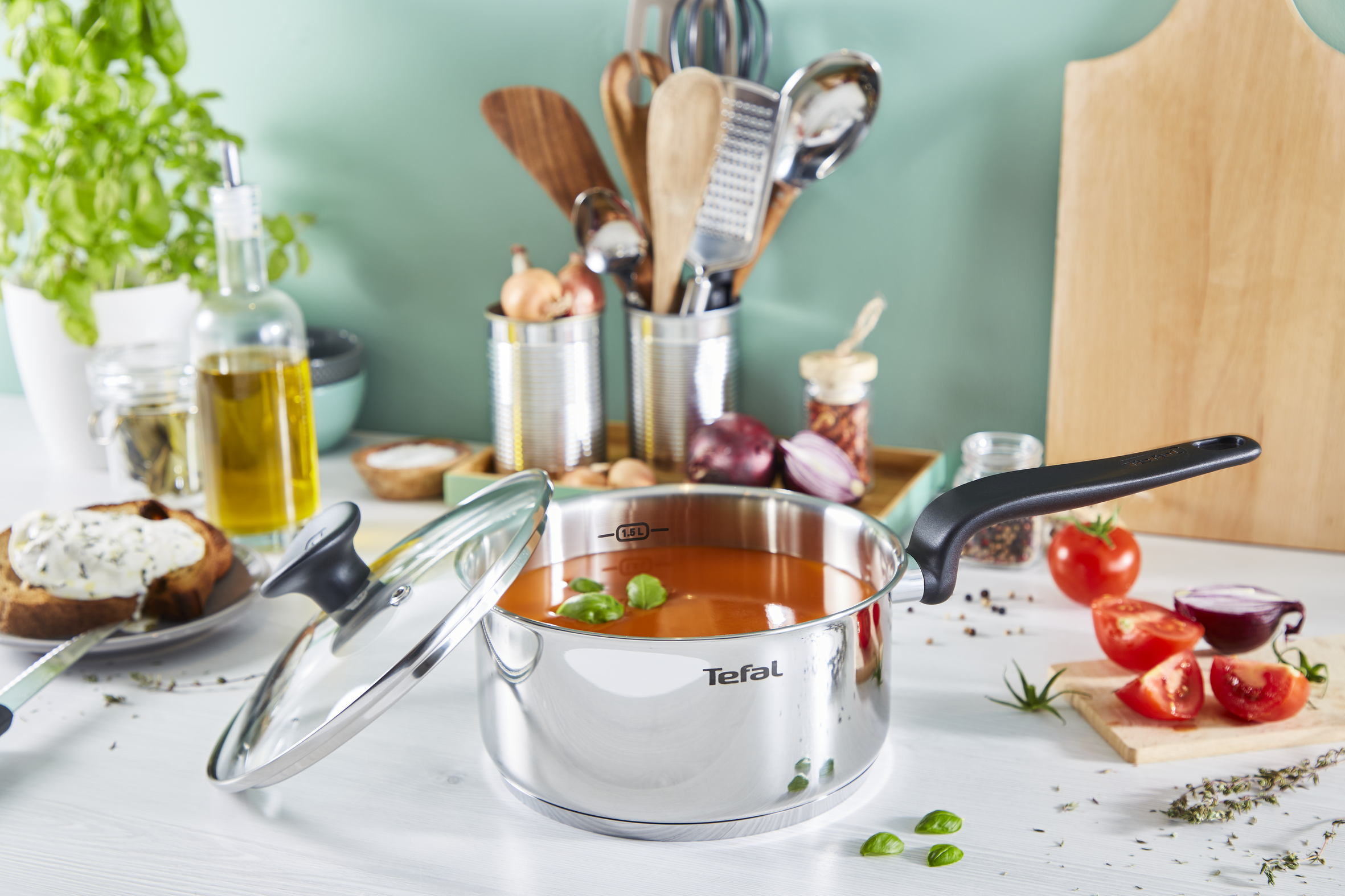 Una olla Tefal brillante con sopa naranja y hojas de albahaca, junto a tomates y cebollas.