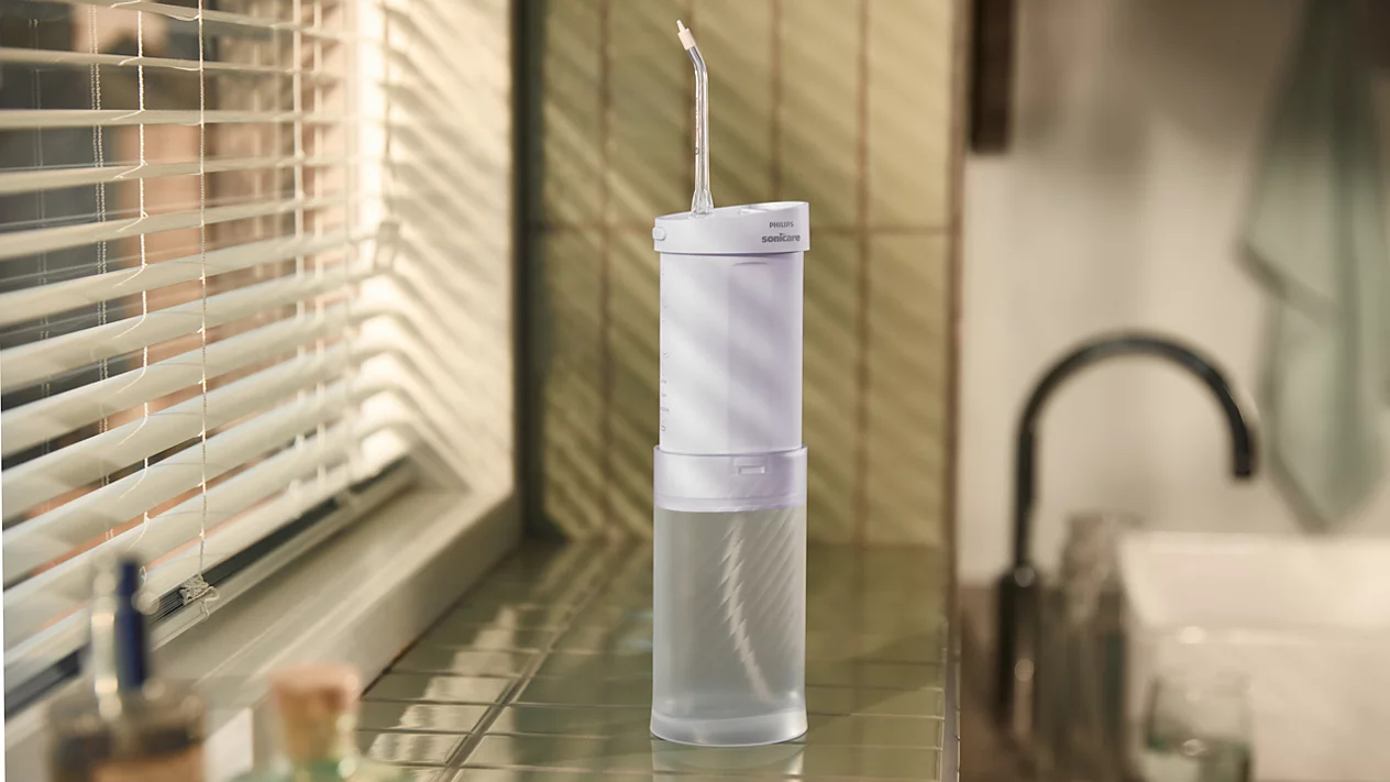 Biała irygator wodny Philips Sonicare na jasnozielonej kafelkowej powierzchni obok okna z żaluzjami.
