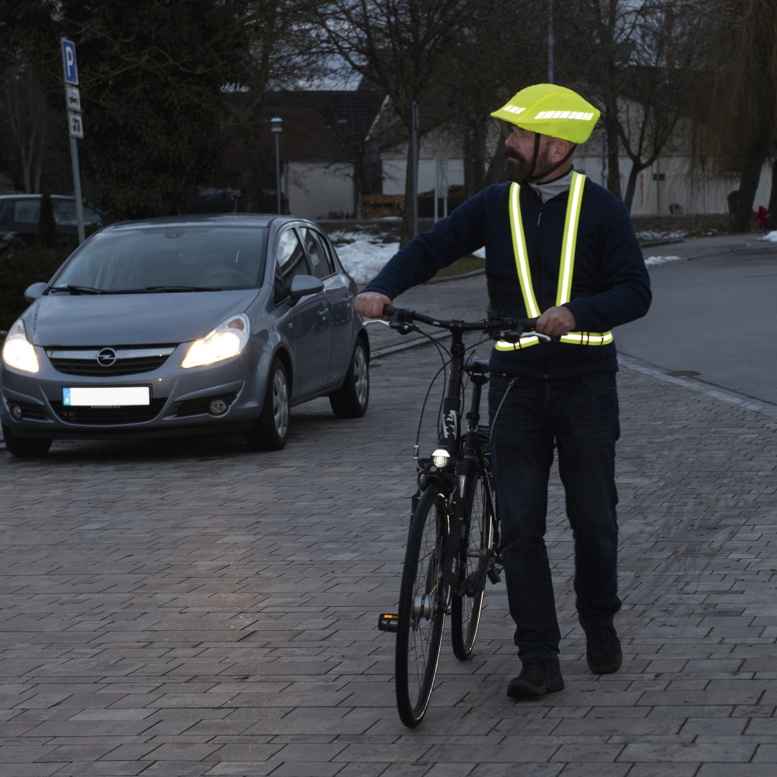 Ein Mann mit Helm und Warnweste schiebt sein Fahrrad neben einem Auto.