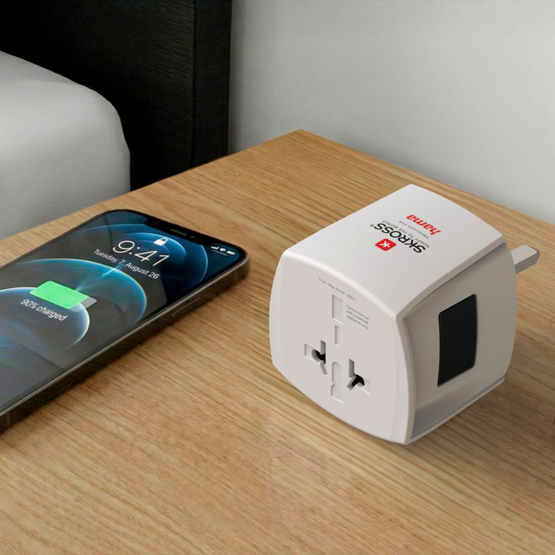 Ein Smartphone, das zu 90% geladen wird, sitzt neben einem weißen Universalreiseadapter auf einem Nachttisch aus Holz.