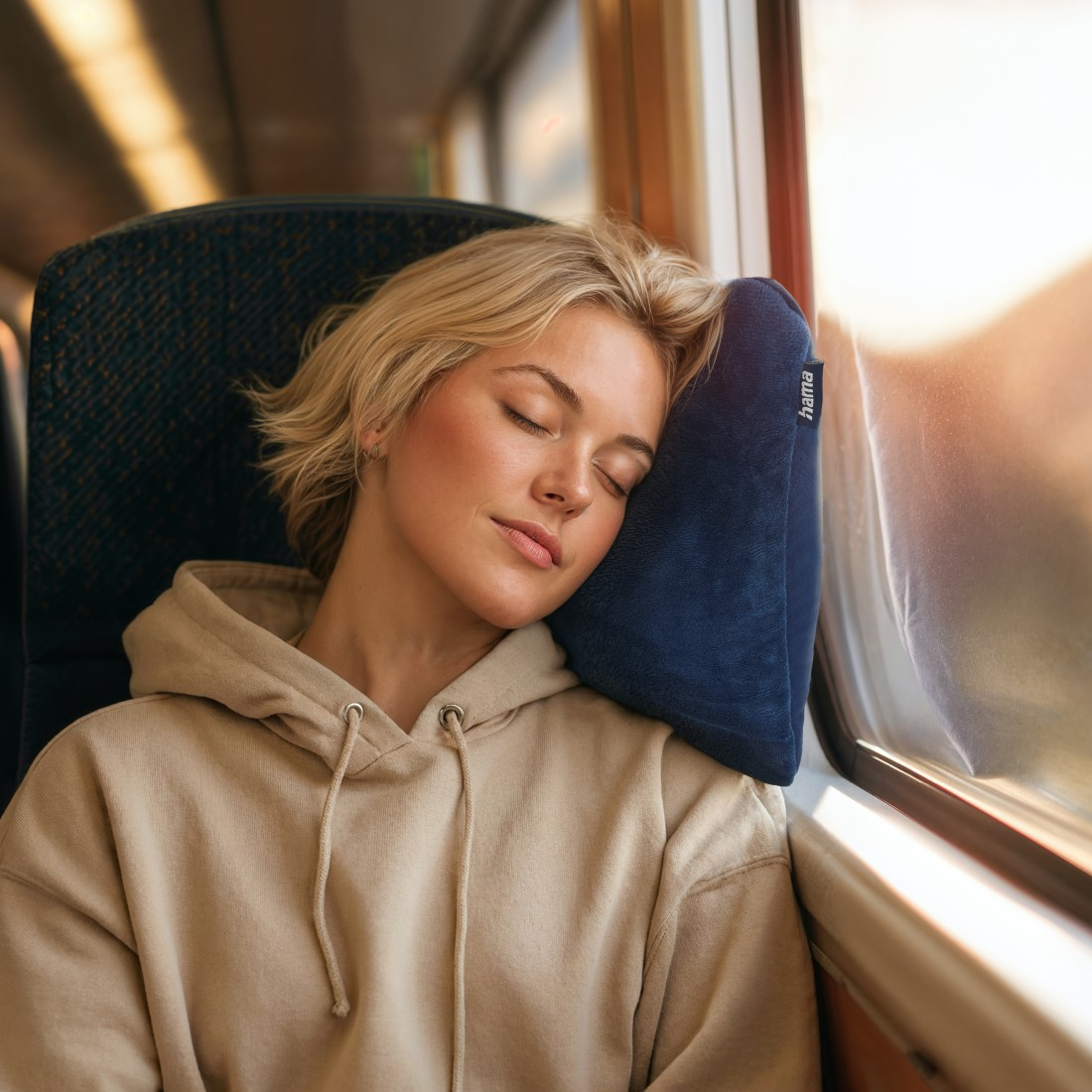Eine Frau mit blonden Haaren schläft auf einem Kissen im Zug. Sie trägt einen beigen Kapuzenpullover.