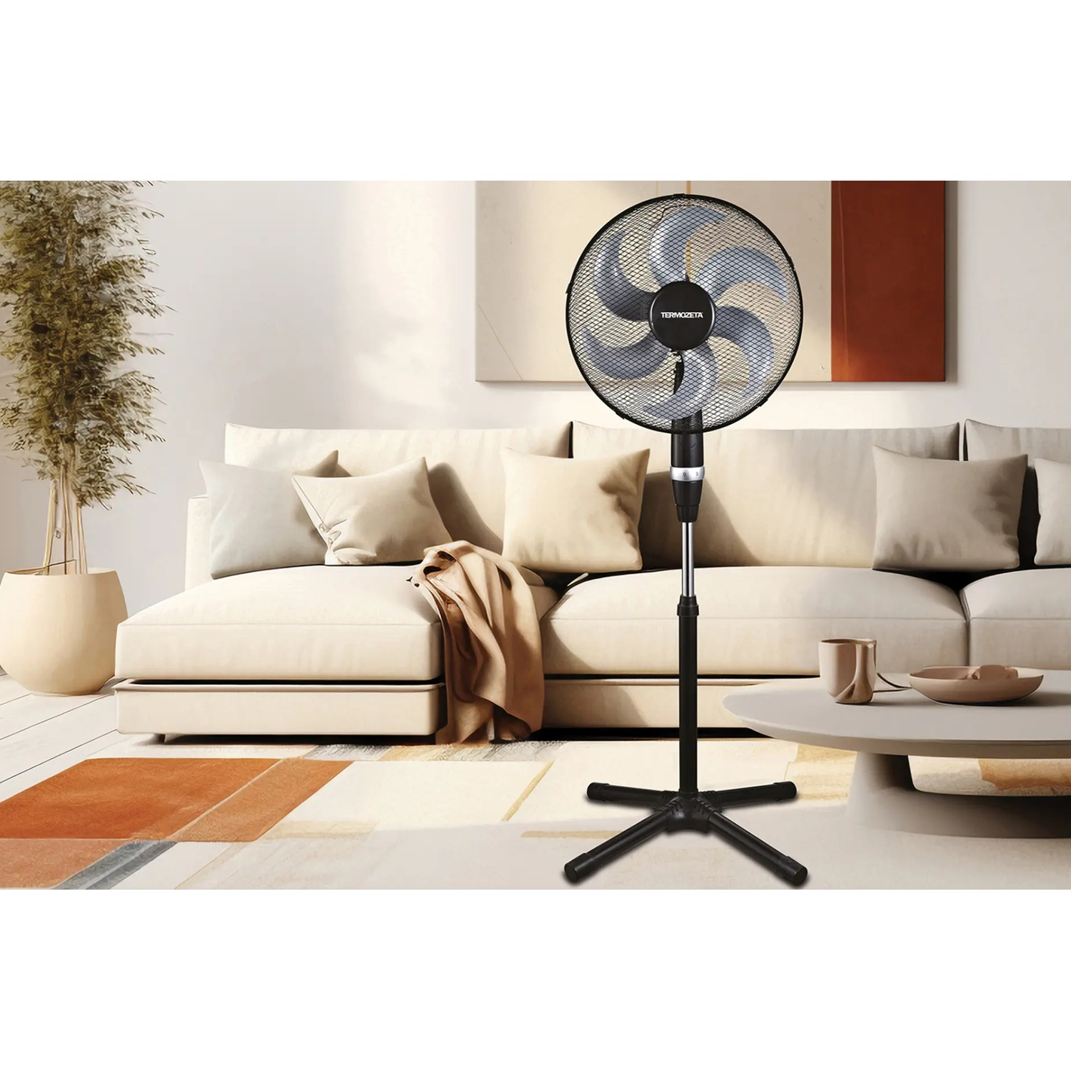 A black standing fan is placed in front of a sofa with cushions, a side table is nearby.