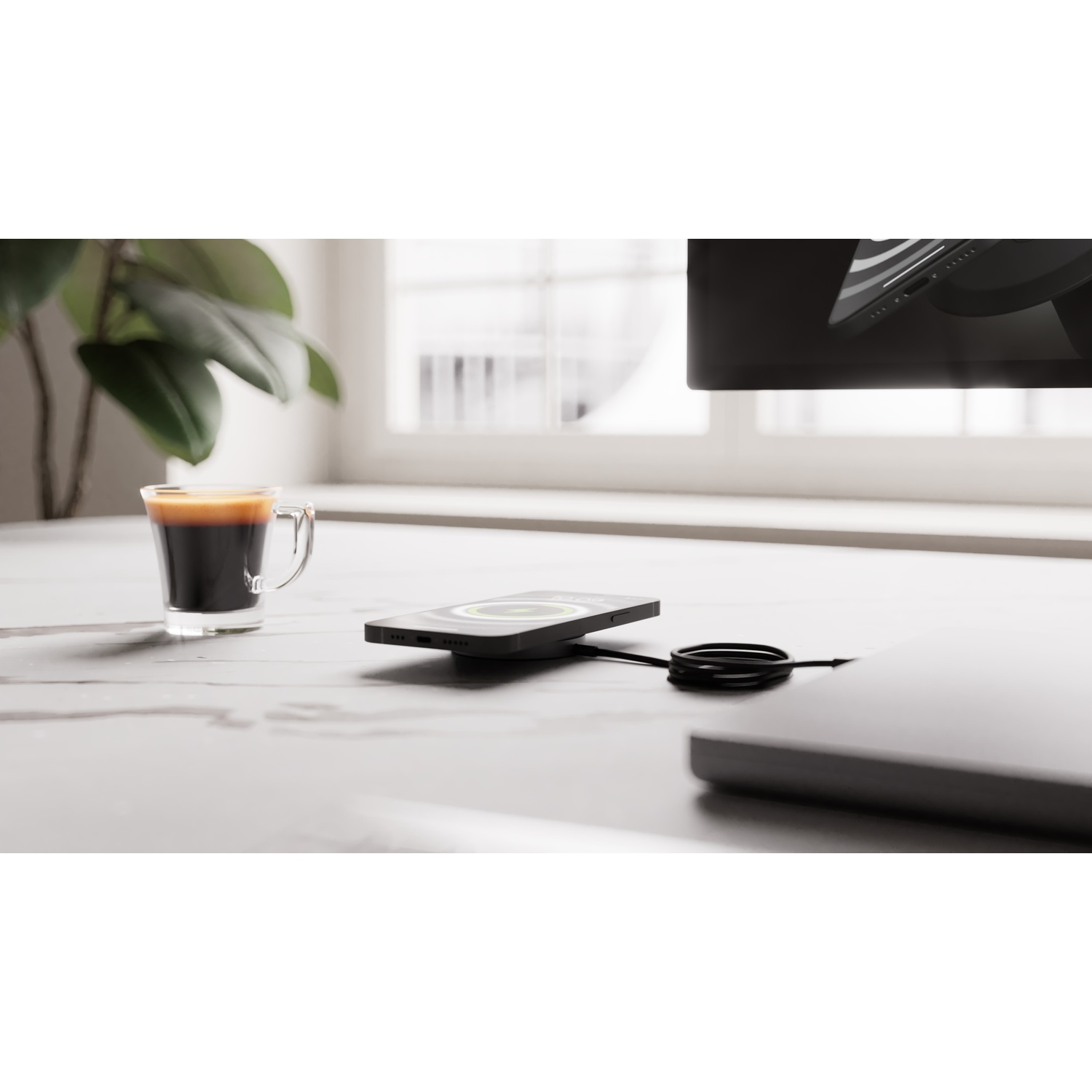 A black smartphone charges on a pad next to a cup of espresso. A plant is nearby.