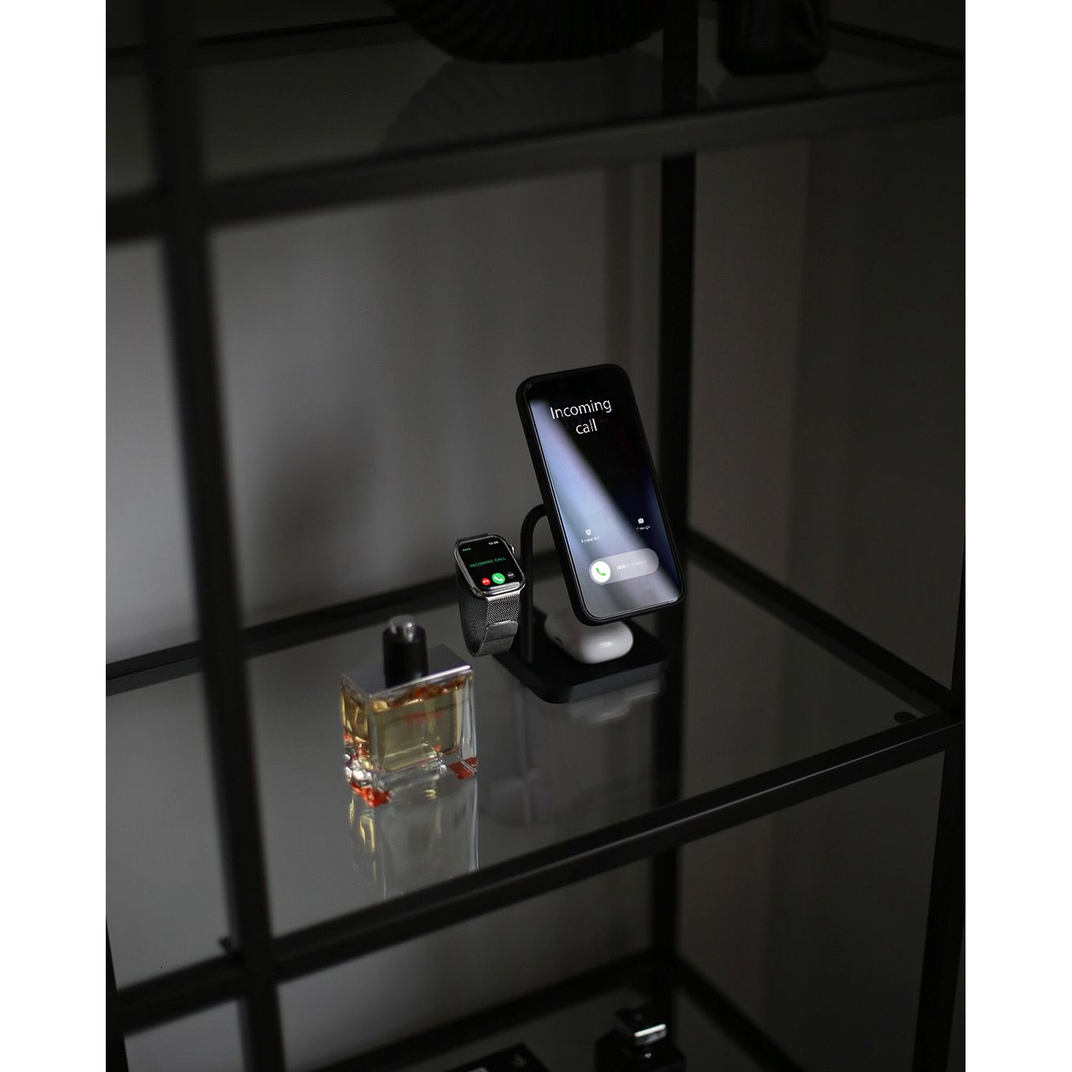 A black smartphone with an incoming call and a smartwatch are displayed on a glass shelf next to a perfume bottle.