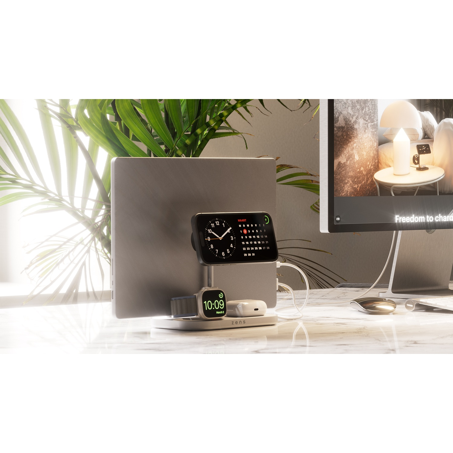 A desk with a charging station holding a phone, smartwatch, and earbuds, next to a laptop and a monitor.