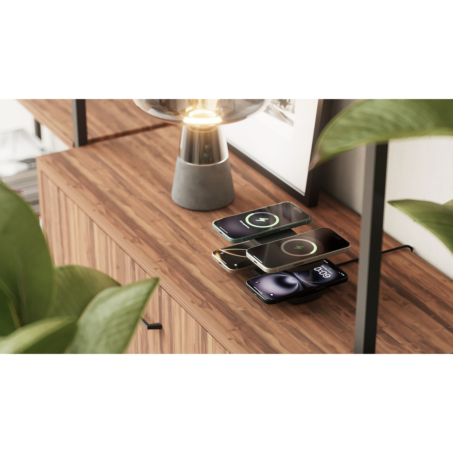 Three smartphones charging wirelessly on a wooden surface next to a lit lamp and a green plant.