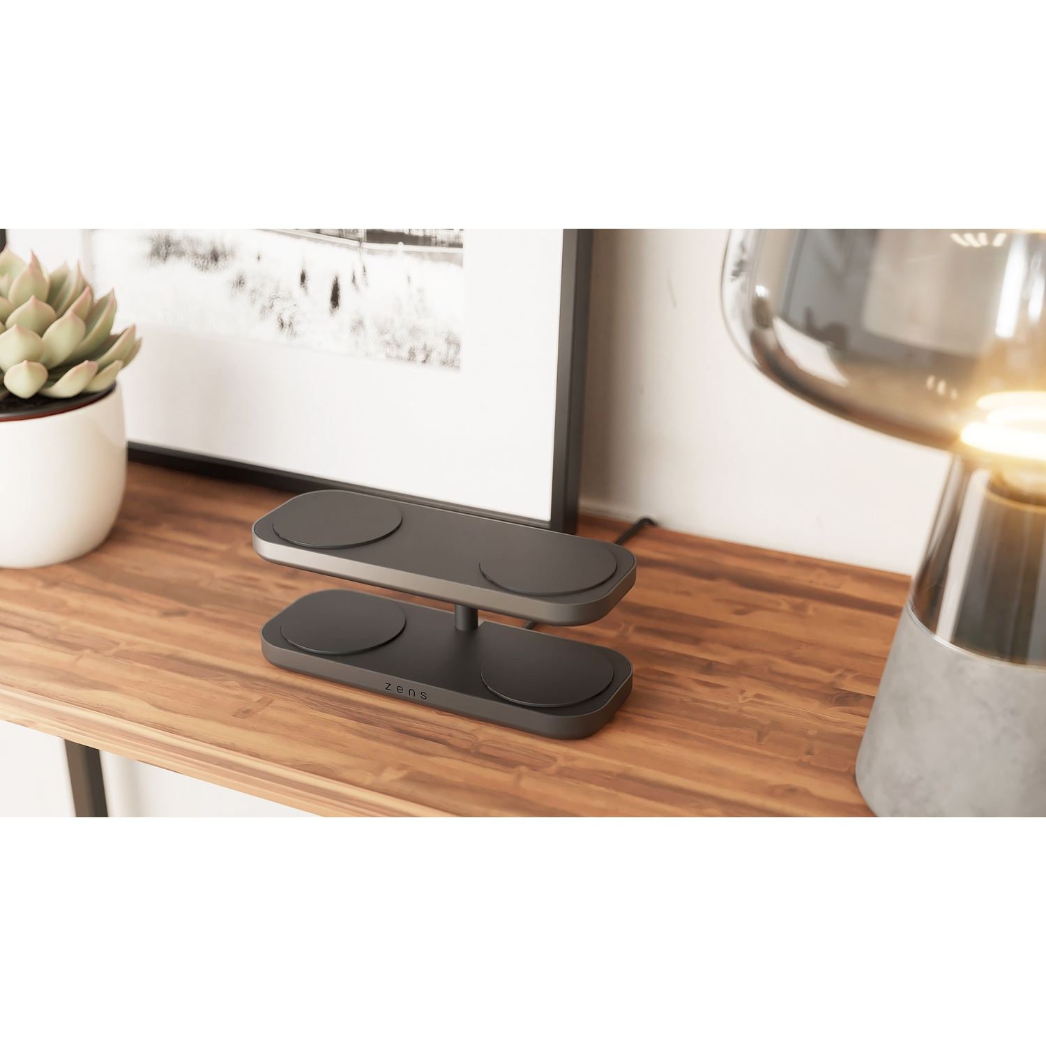 A black Zens wireless charger, with two tiers and circular charging pads, sits on a wooden table next to a succulent plant and a lamp.