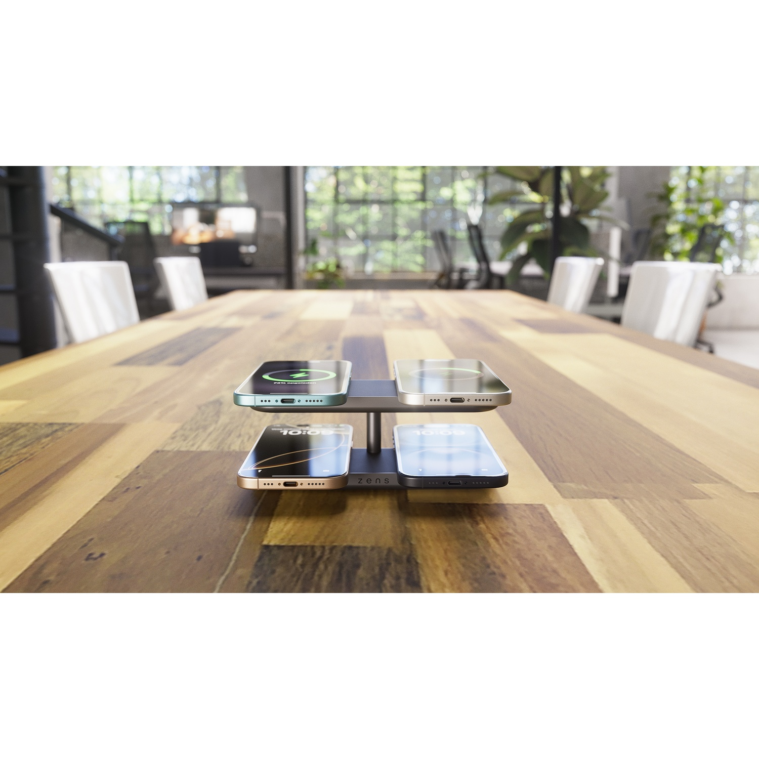 Four smartphones on a charging stand on a wooden table in a bright office.