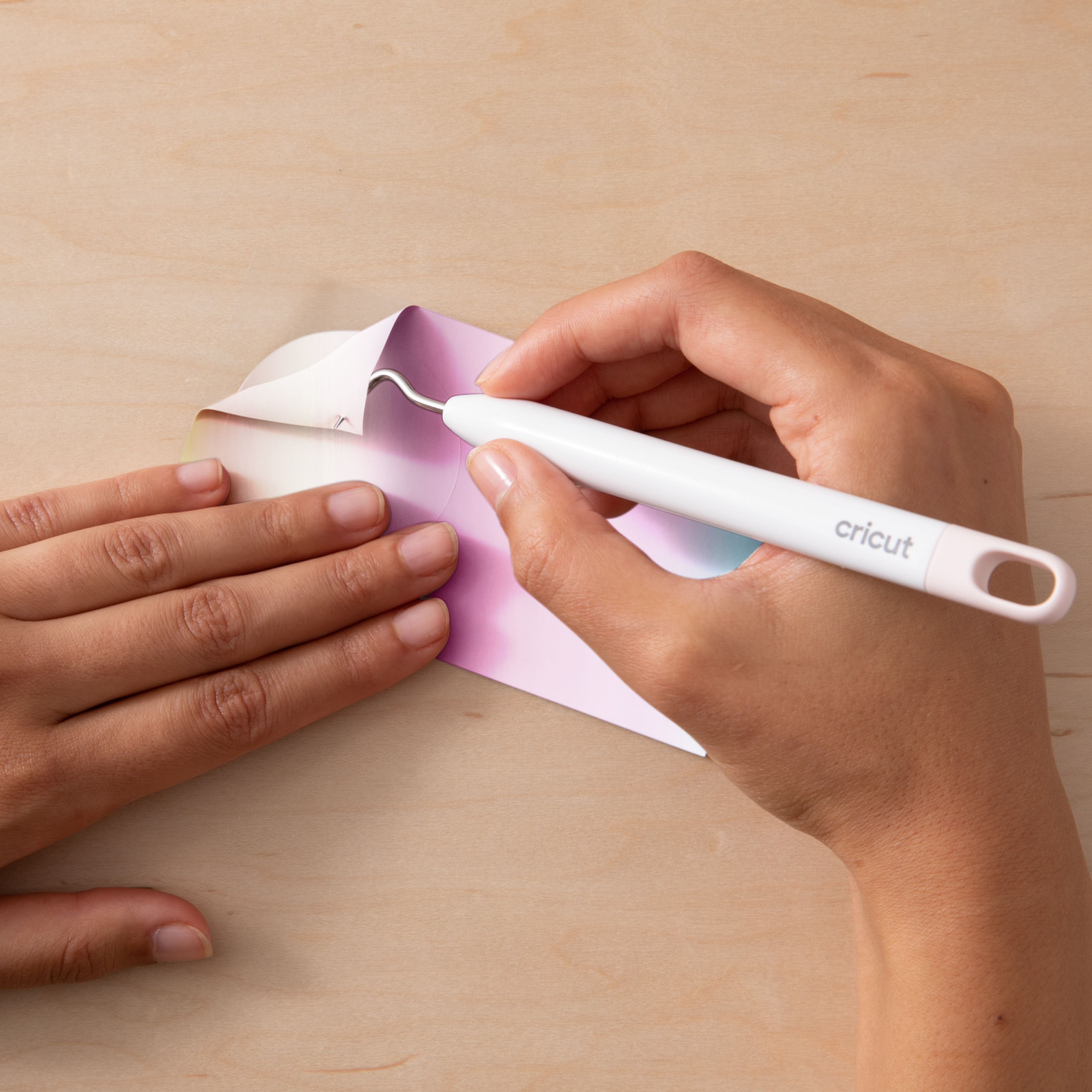 Eller ahşap üzerinde mor/pembe kağıt ve Cricut ayıklama aleti tutuyor.